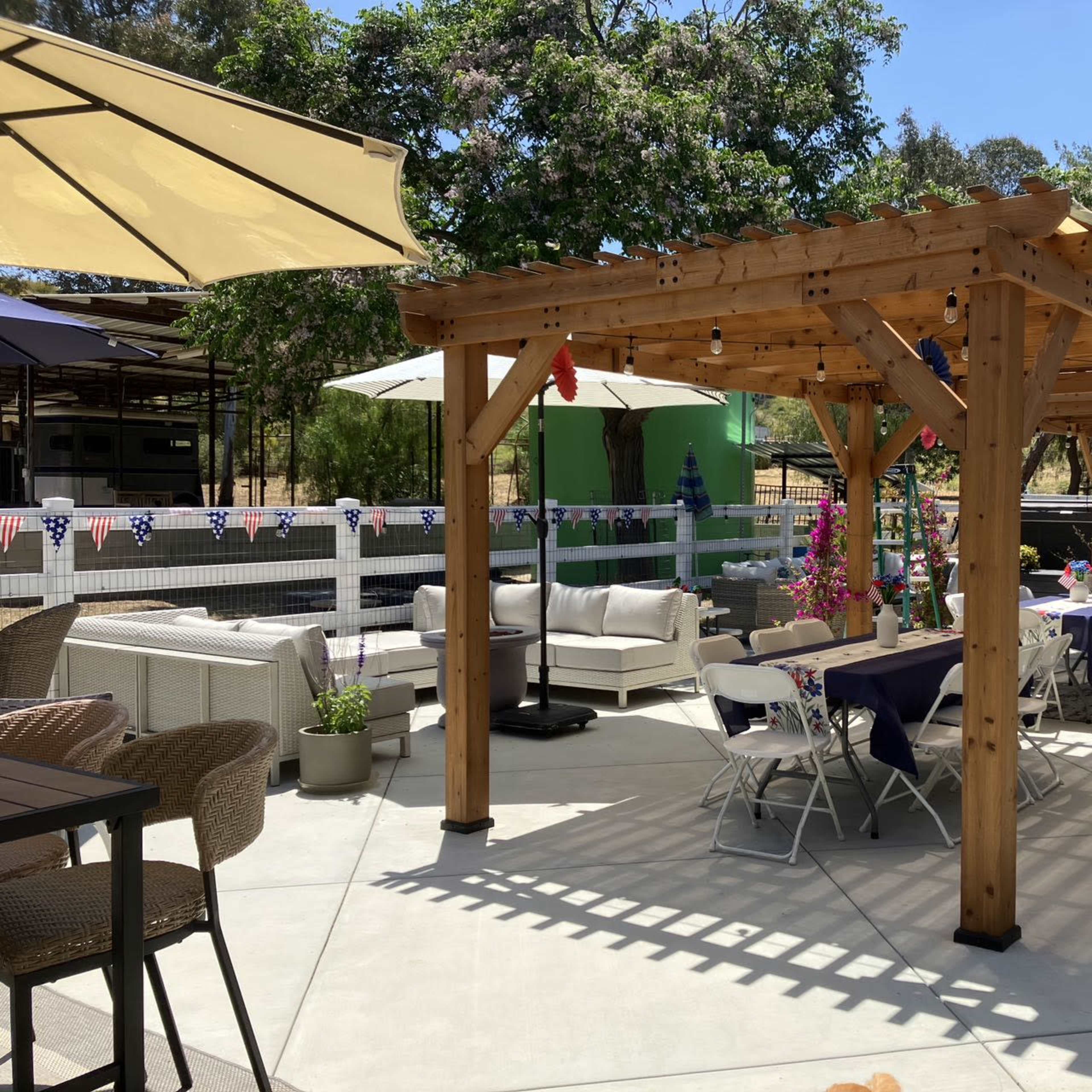 A wooden pergola with tables and chairs is set up in a patio area surrounded by greenery and decorative banners.