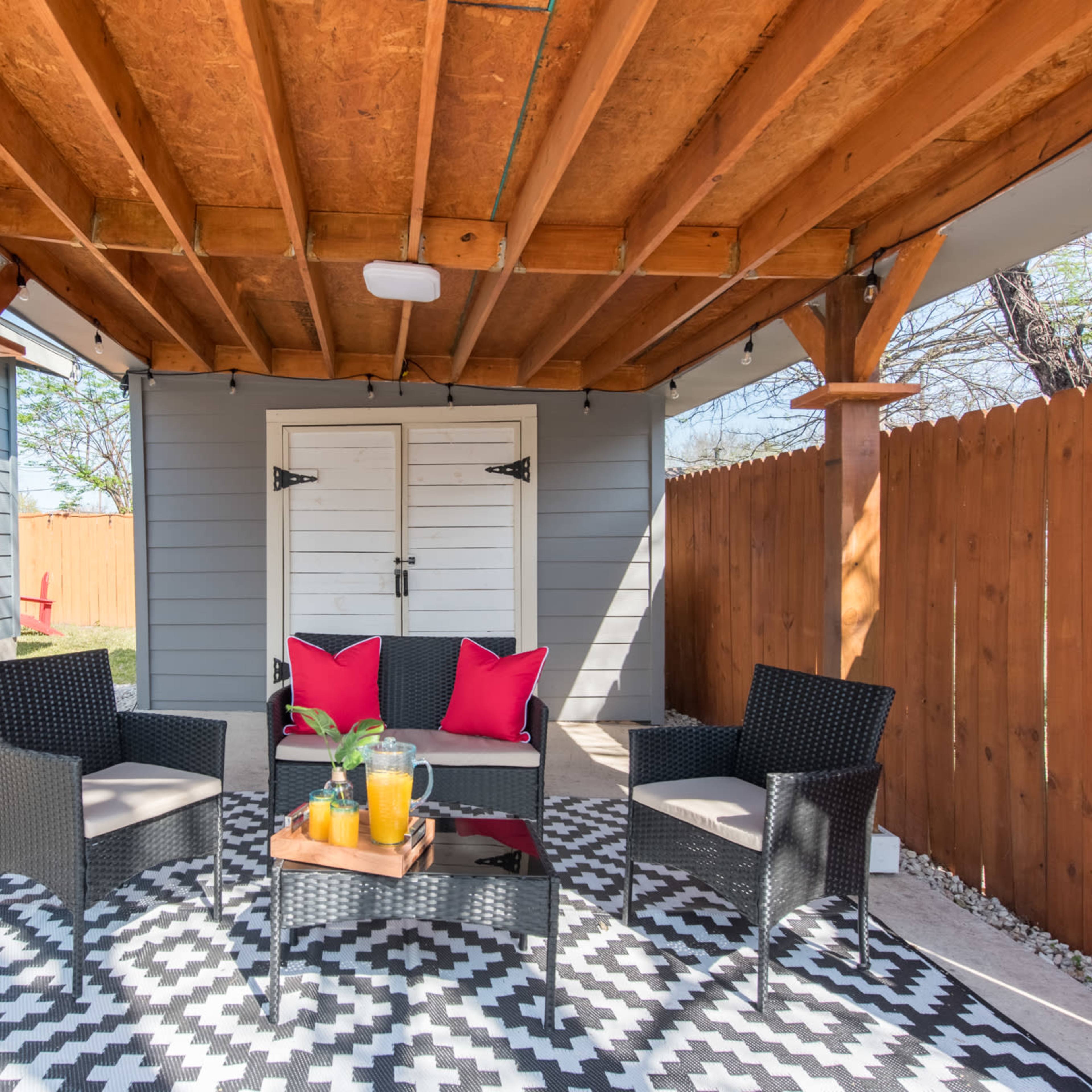 A covered outdoor seating area with four chairs arranged around a small table, set on a patterned rug beneath wooden beams.
