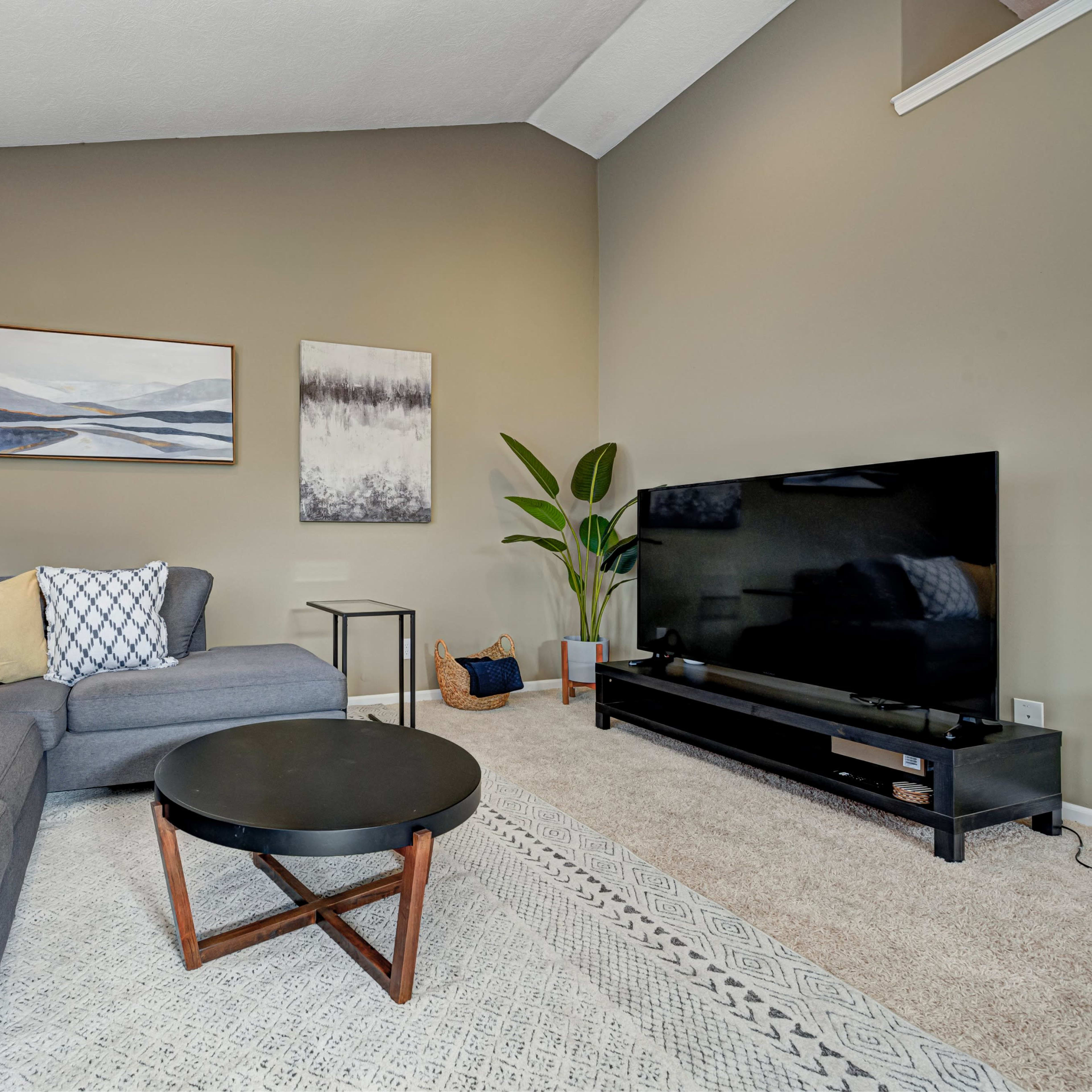 A living room with a sectional sofa, a round coffee table, a television stand, and two framed artworks on the walls.