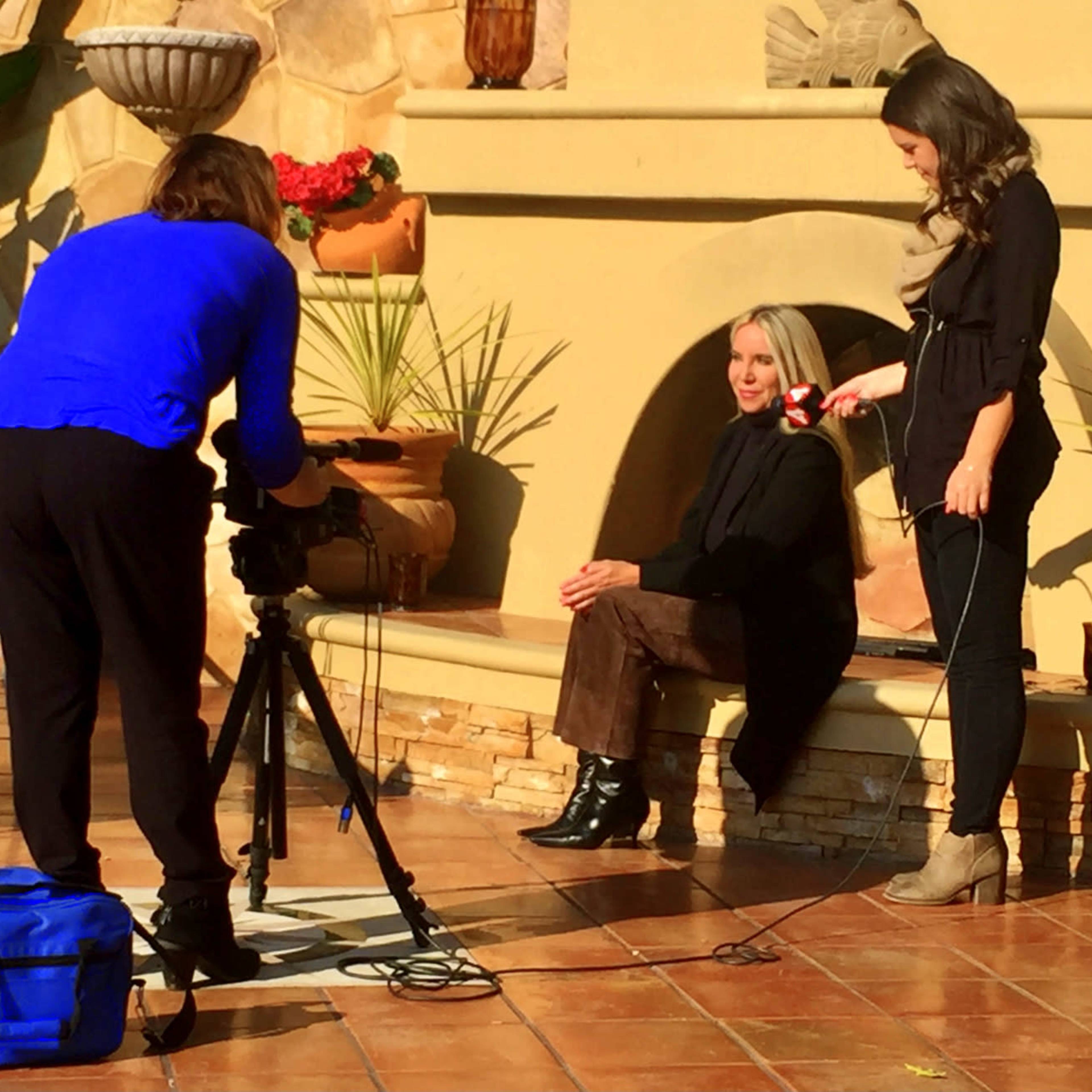 A woman sits against a fireplace while another woman adjusts a microphone, and a third person sets up a camera on a tripod.