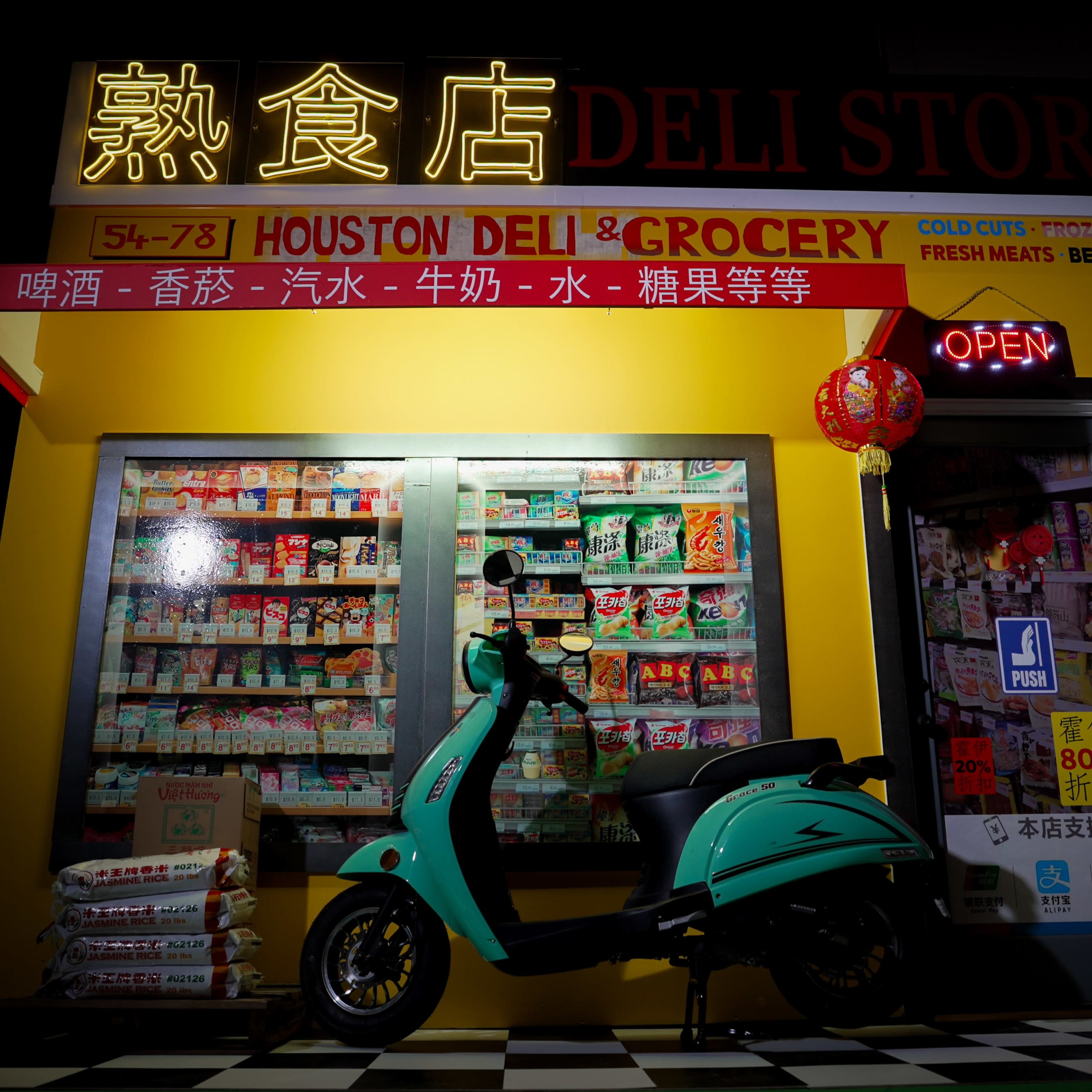 A brightly lit deli store featuring a turquoise scooter in front of shelves stocked with various groceries and a red lantern hanging above the entrance.