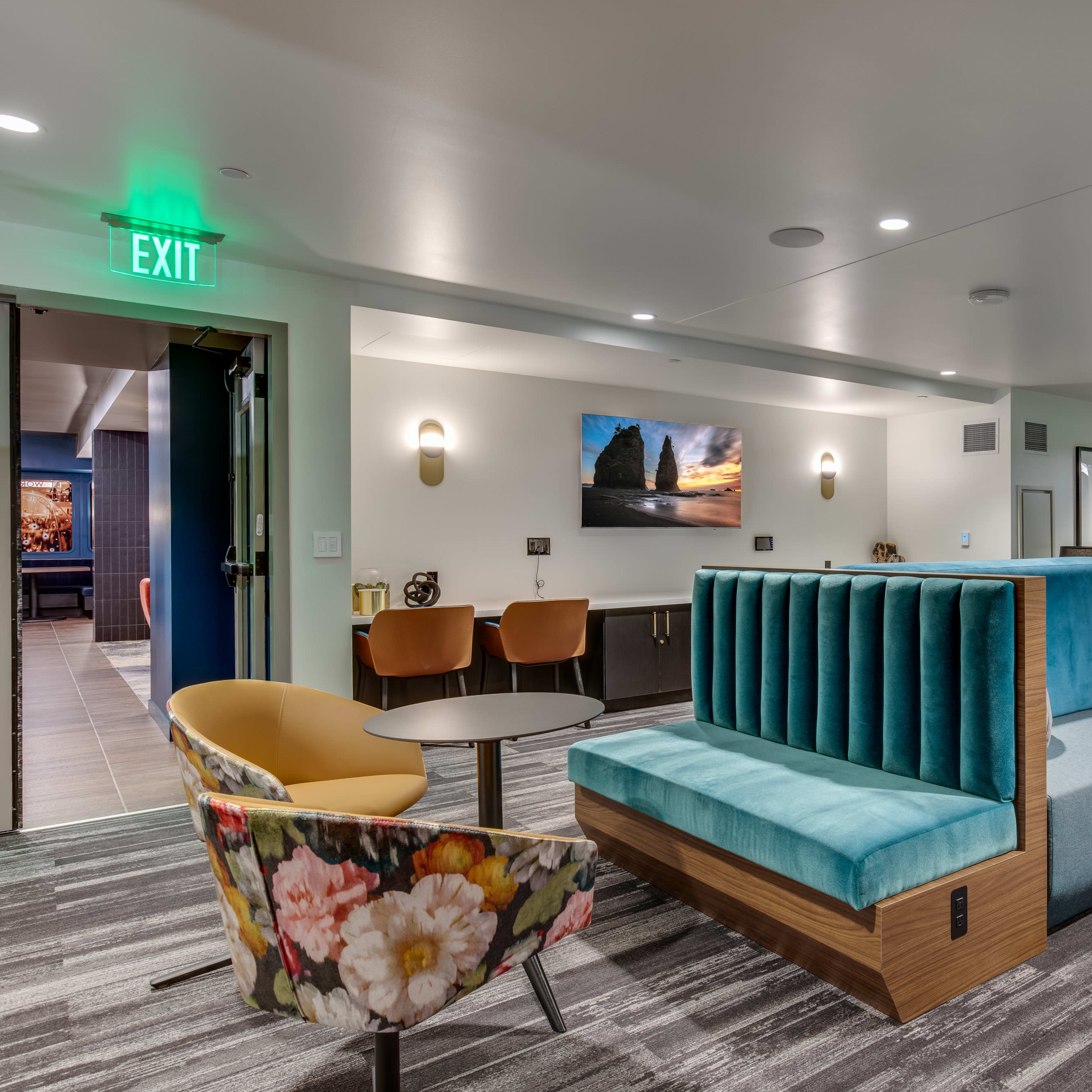 A modern lounge area featuring a large turquoise sectional sofa, a table, and colorful chairs against a backdrop of muted walls and patterned flooring.