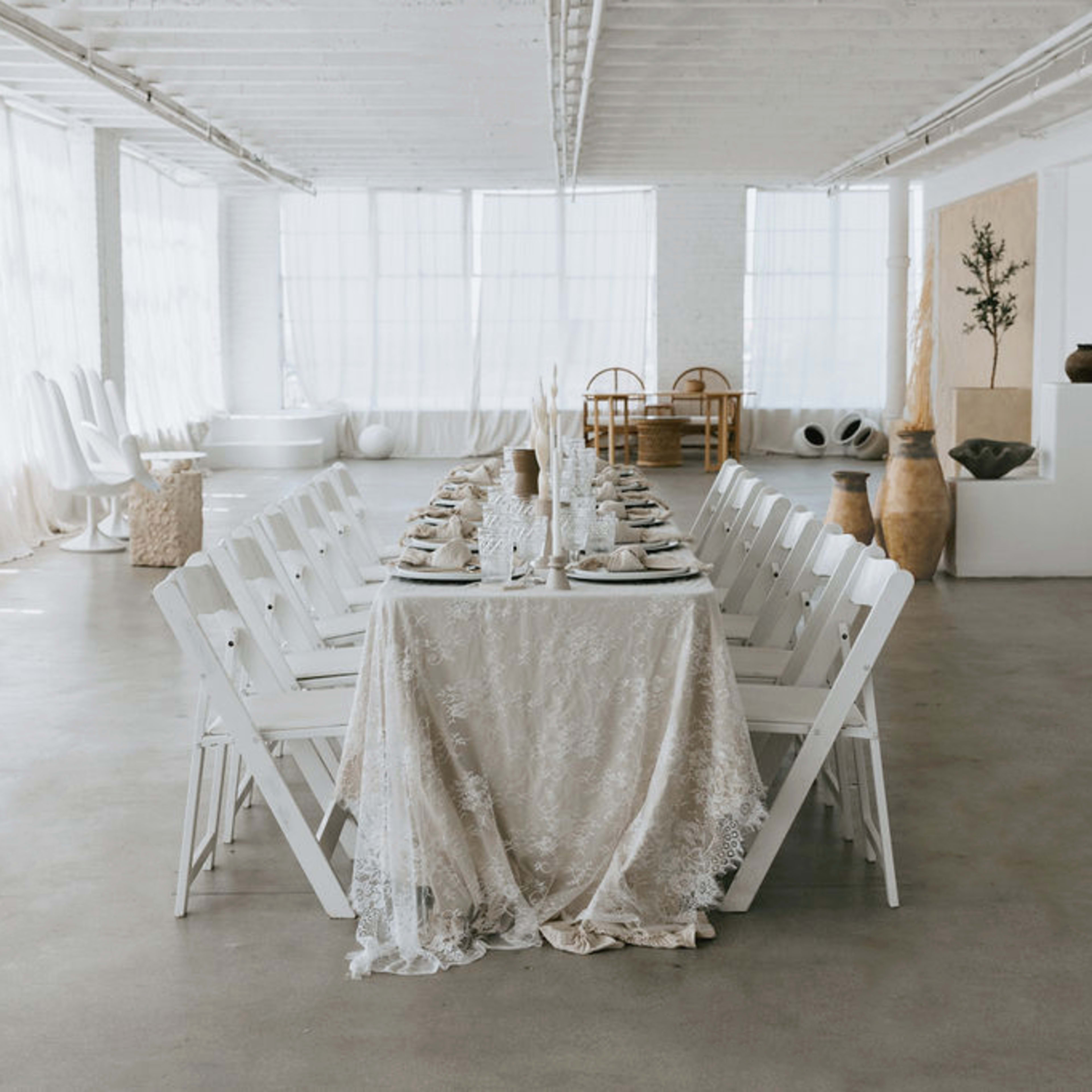 A long, elegantly set dining table with white chairs is arranged in a bright, airy room featuring large windows and minimalistic decor.