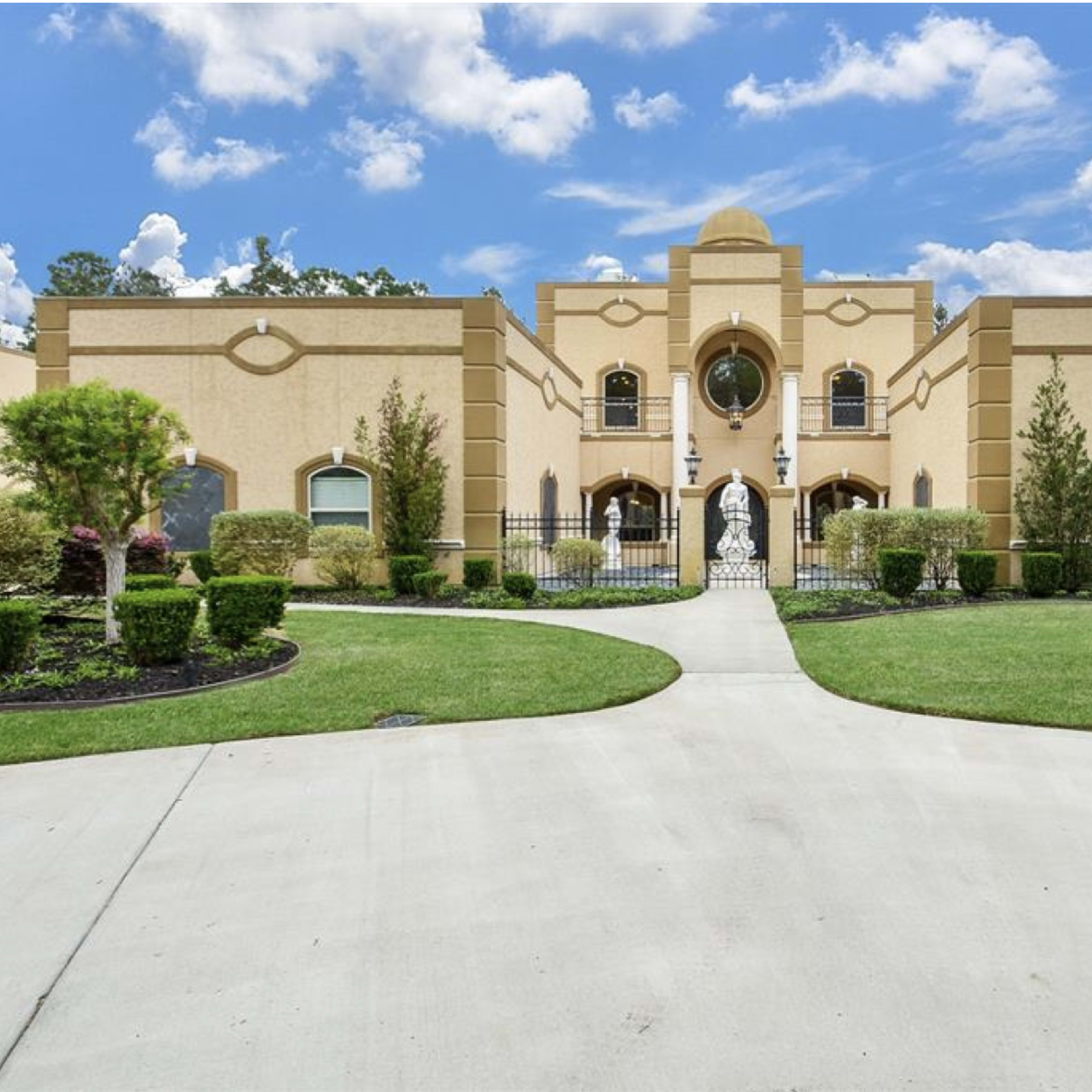 A large, ornate house framed by neatly trimmed shrubs and a paved pathway leading to a central entrance with a statue.