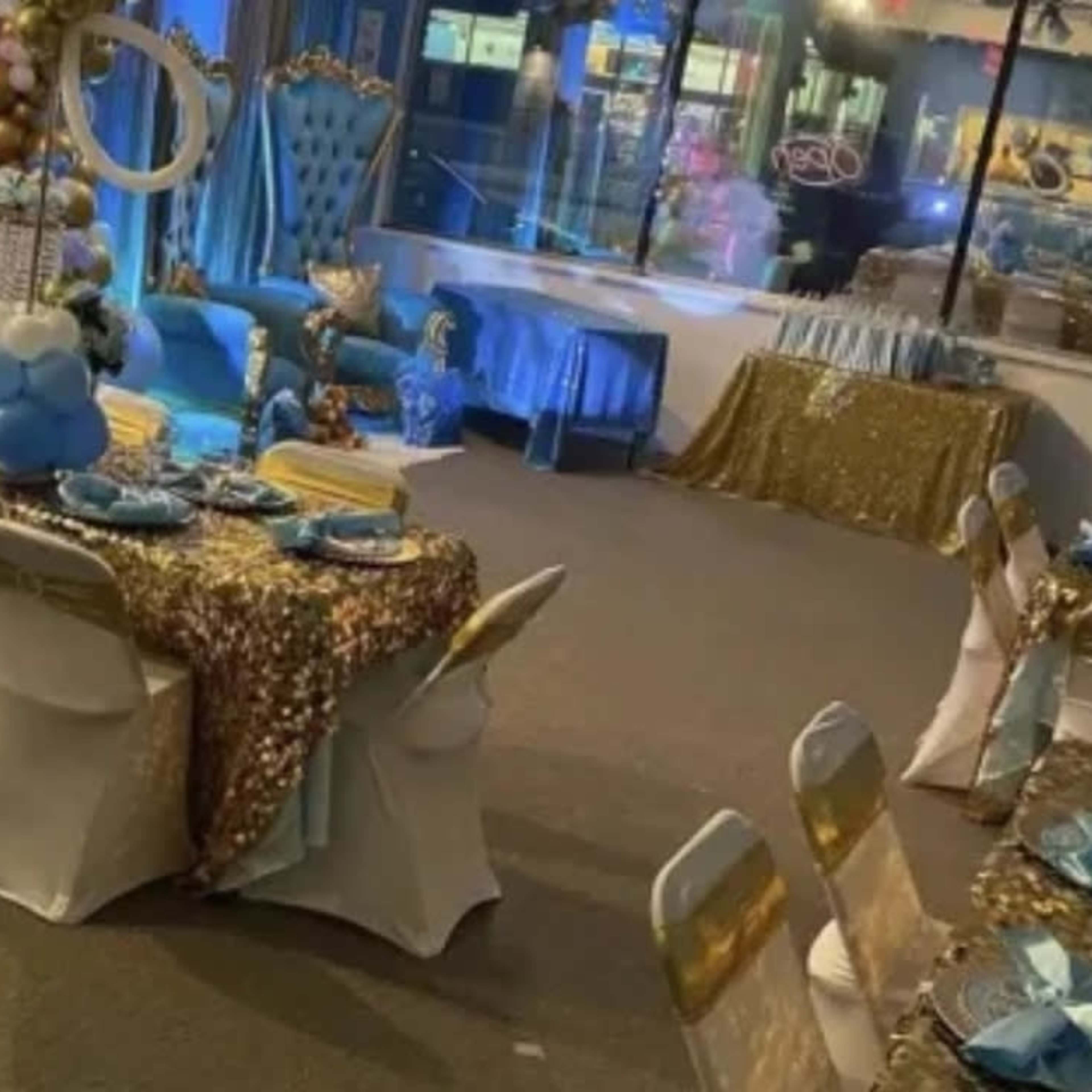 A decorated party venue featuring tables with sequined tablecloths, blue and gold accents, and a large chair at one end.