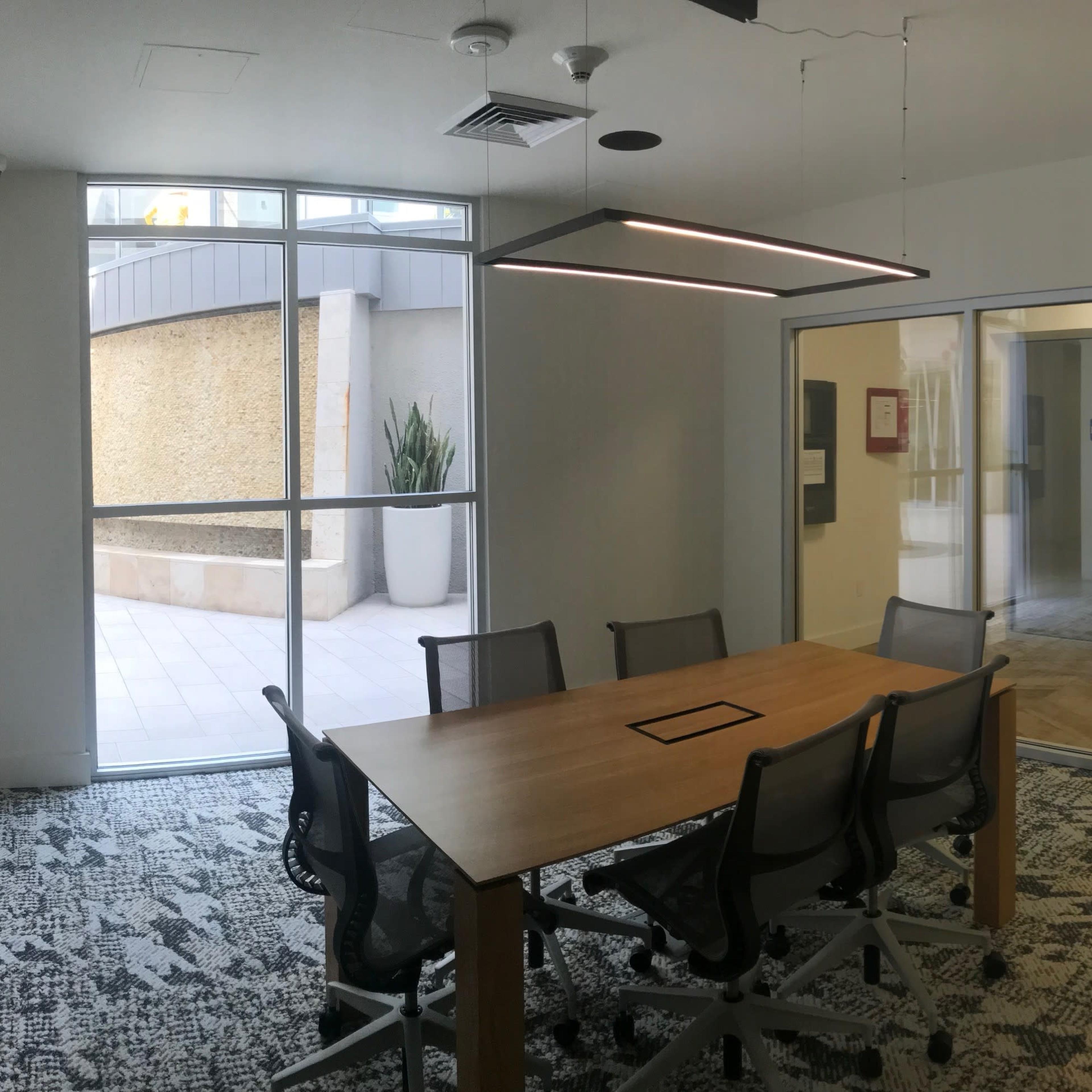 A modern conference room features a large wooden table surrounded by rolling chairs and glass walls that open to an outdoor area.