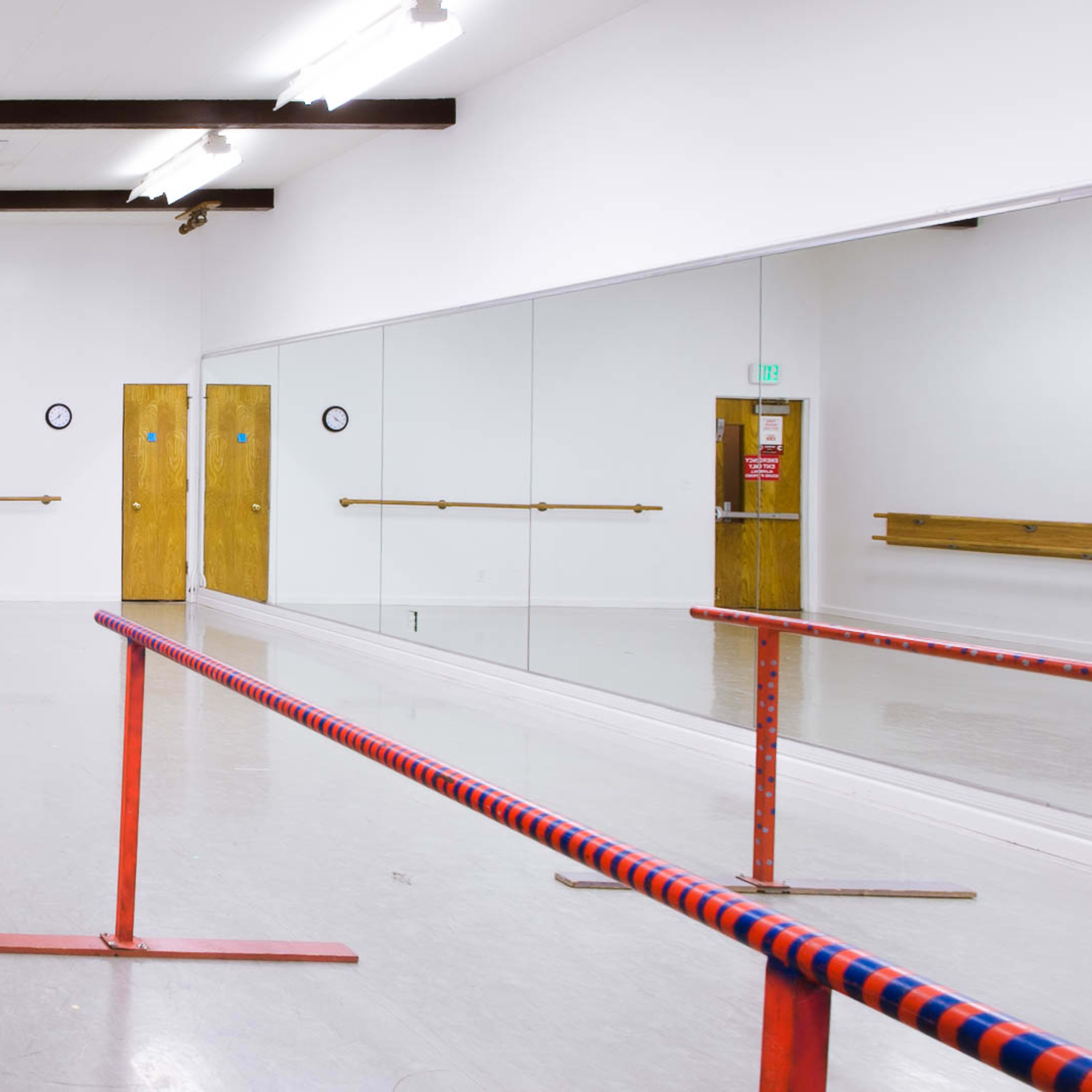 A bright dance studio features a mirrored wall, wooden barre, and double doors with exit signs.