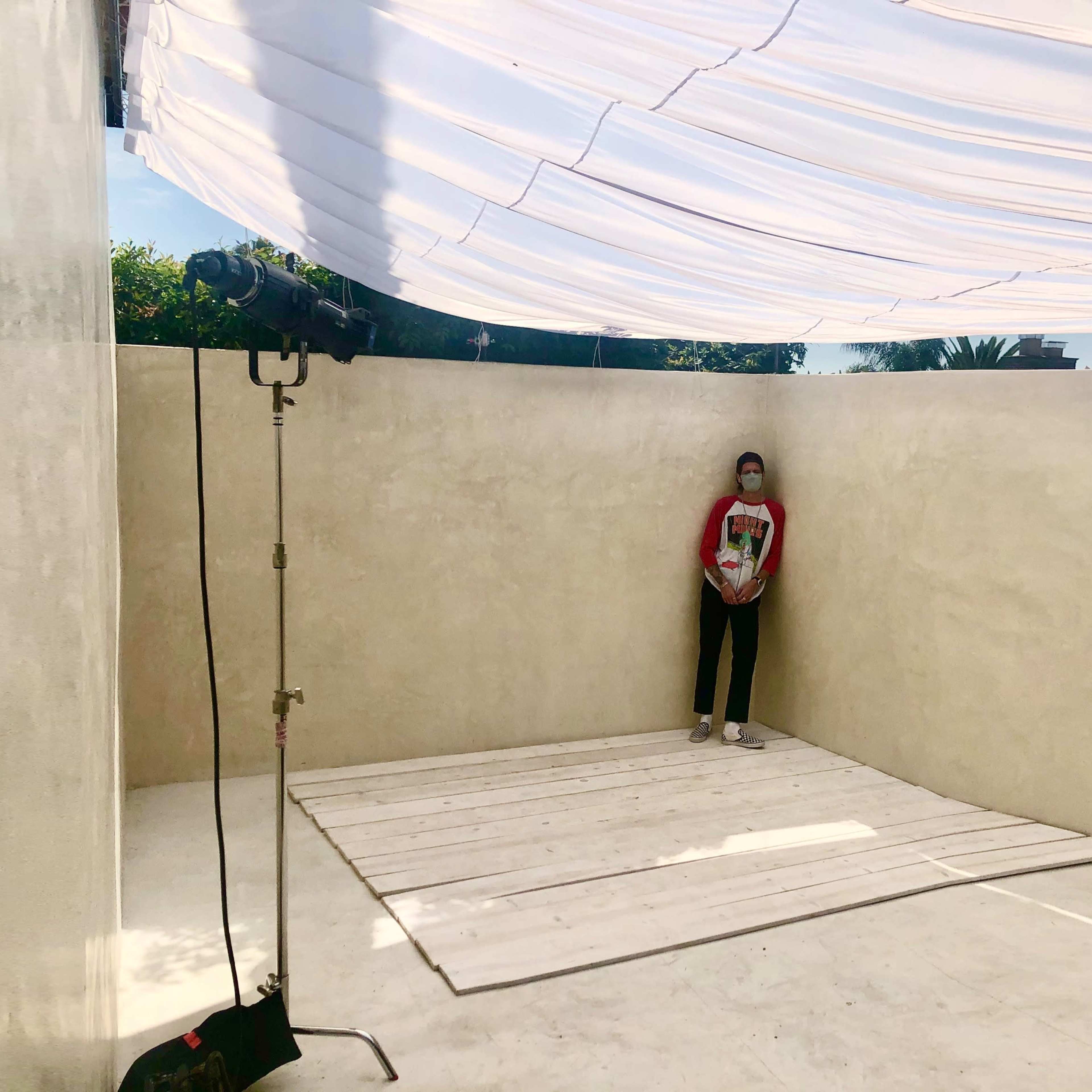 A person stands on a wooden platform in a minimally decorated outdoor space with a fabric canopy overhead.
