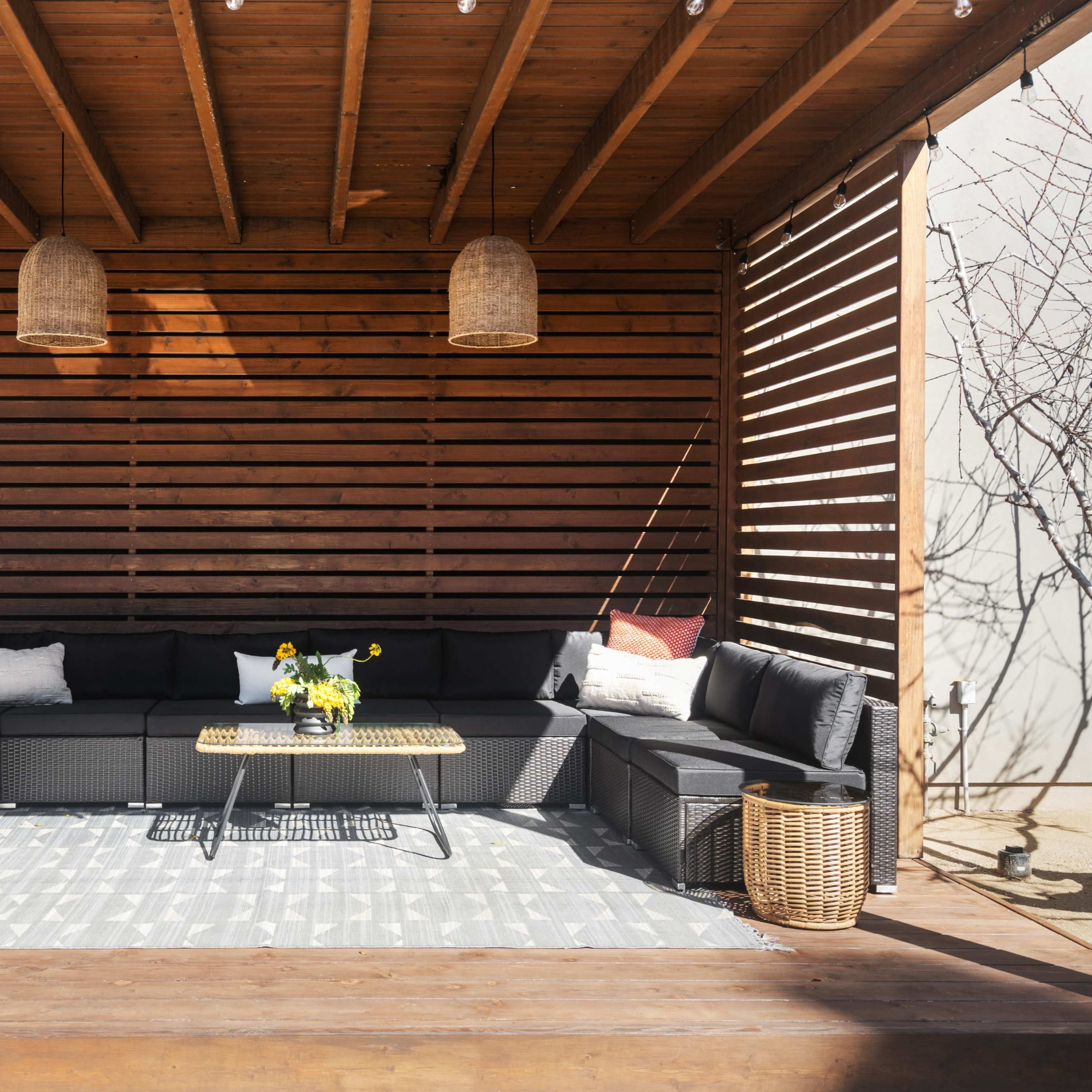 A modern outdoor seating area features a black sectional sofa with decorative pillows, a coffee table, and a patterned rug under wooden beams with two hanging light fixtures.