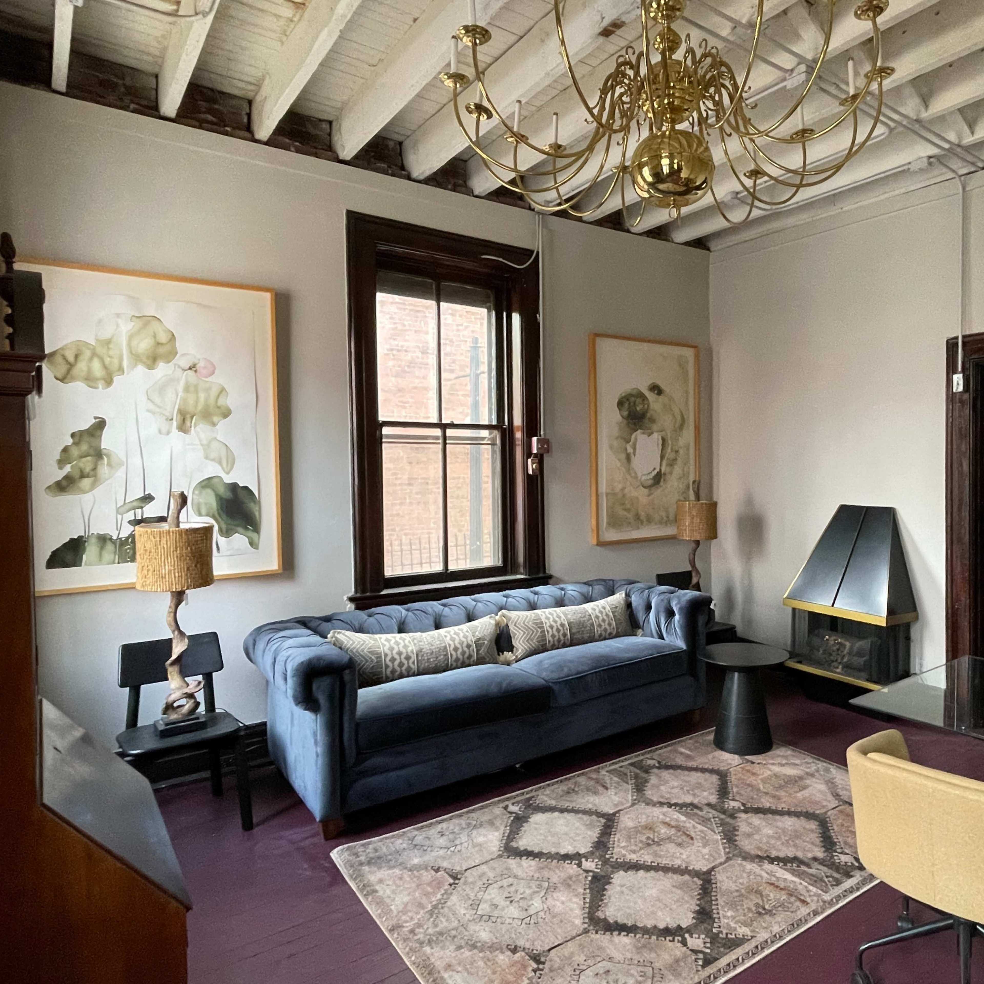 A living room featuring a blue couch, a glass dining table, and artwork on the walls, with a chandelier hanging from the ceiling.