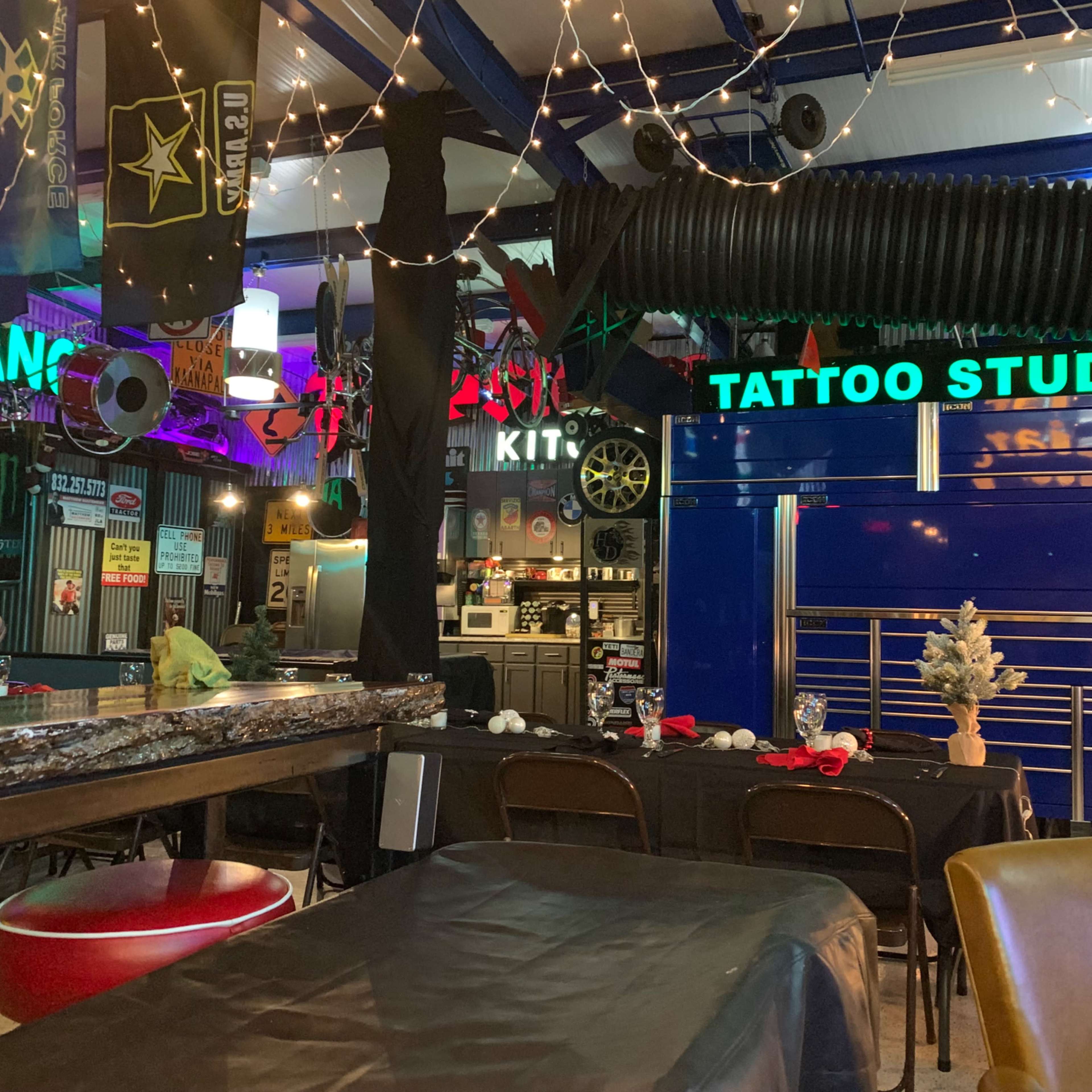 An indoor space featuring a bar area, a tattoo studio sign, and various colorful decorations, with tables set for dining.