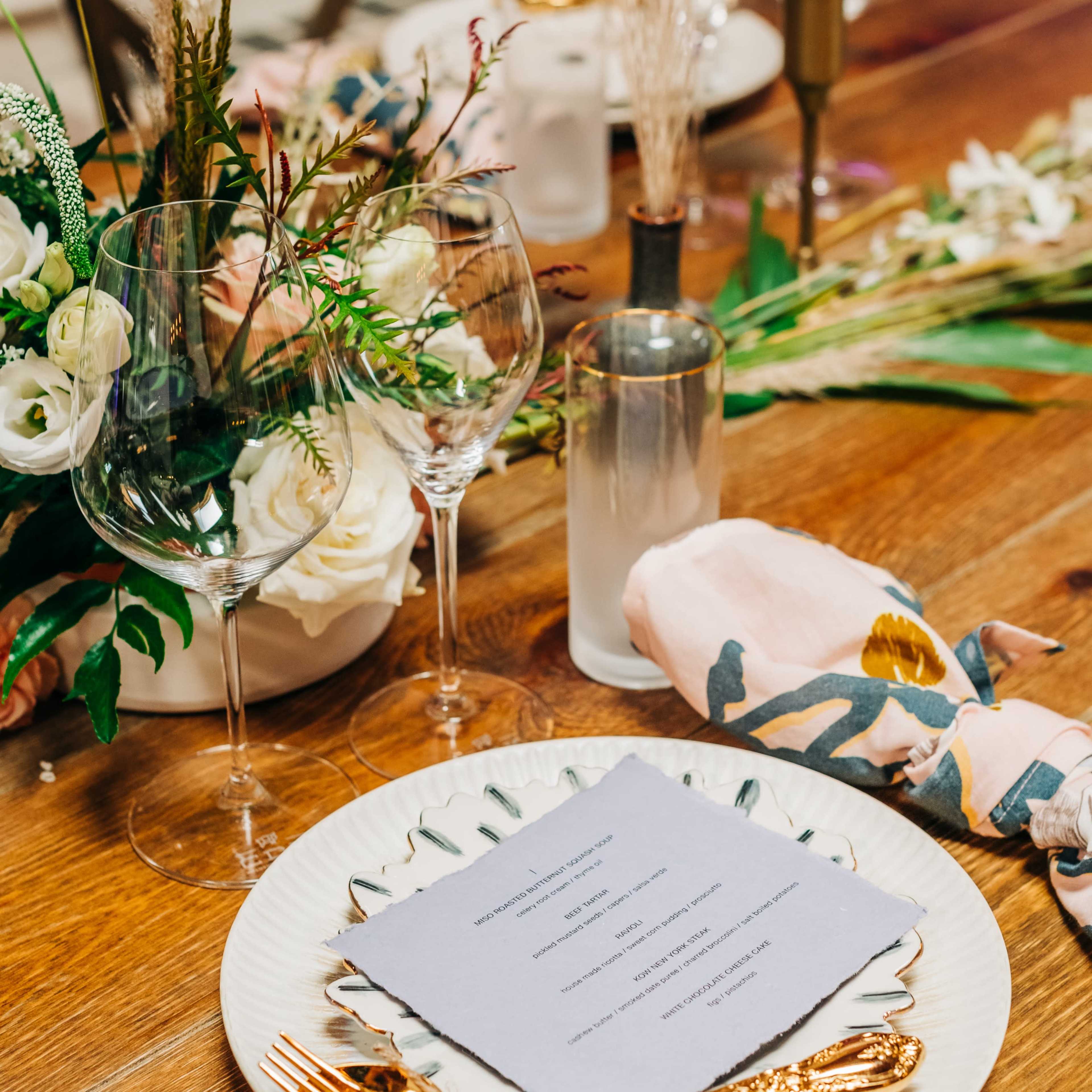 A beautifully set dining table features a floral centerpiece, elegant glassware, golden utensils, and a menu card atop a decorative plate.