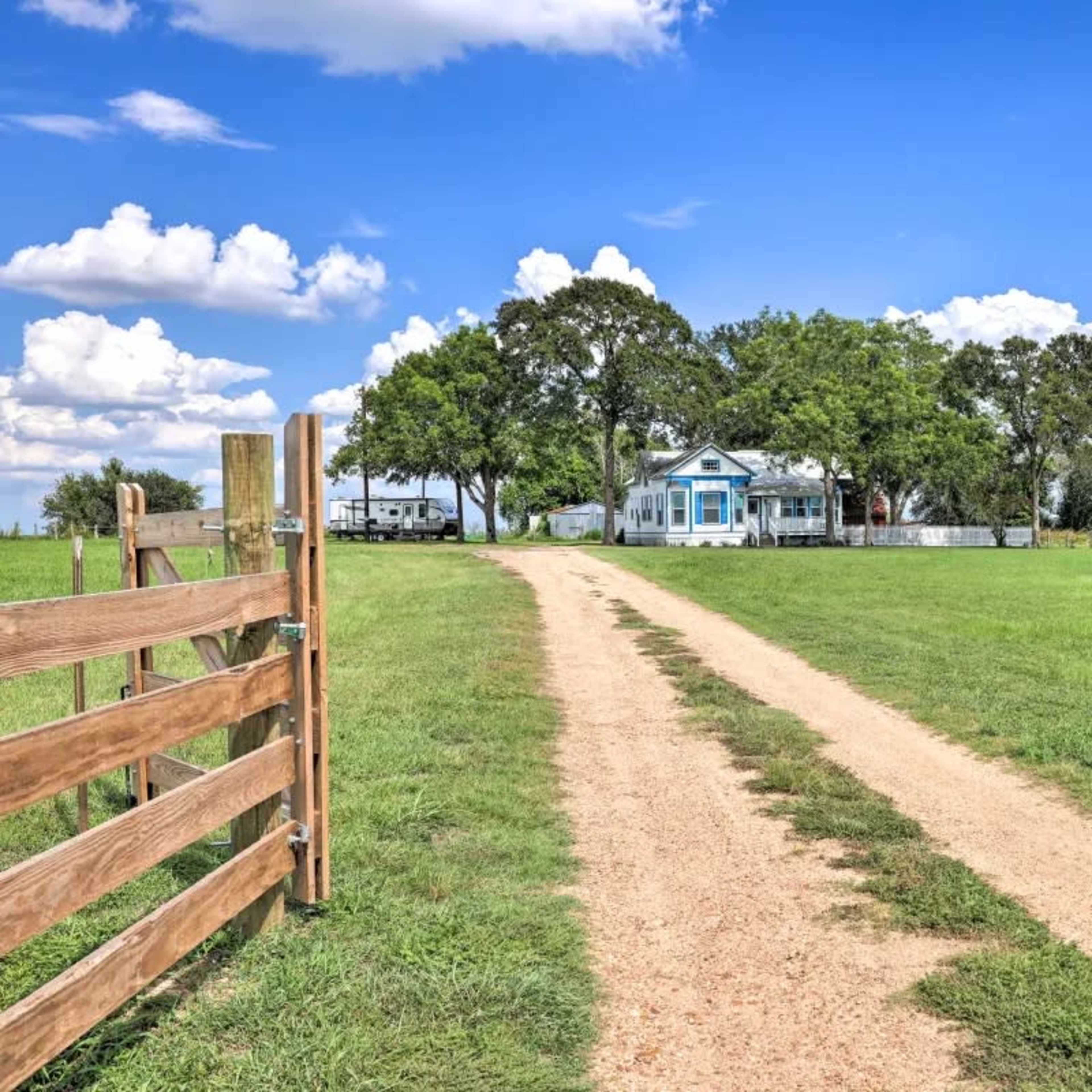 A dirt driveway leads from a wooden gate towards a house surrounded by trees and open grassland under a blue sky with clouds.
