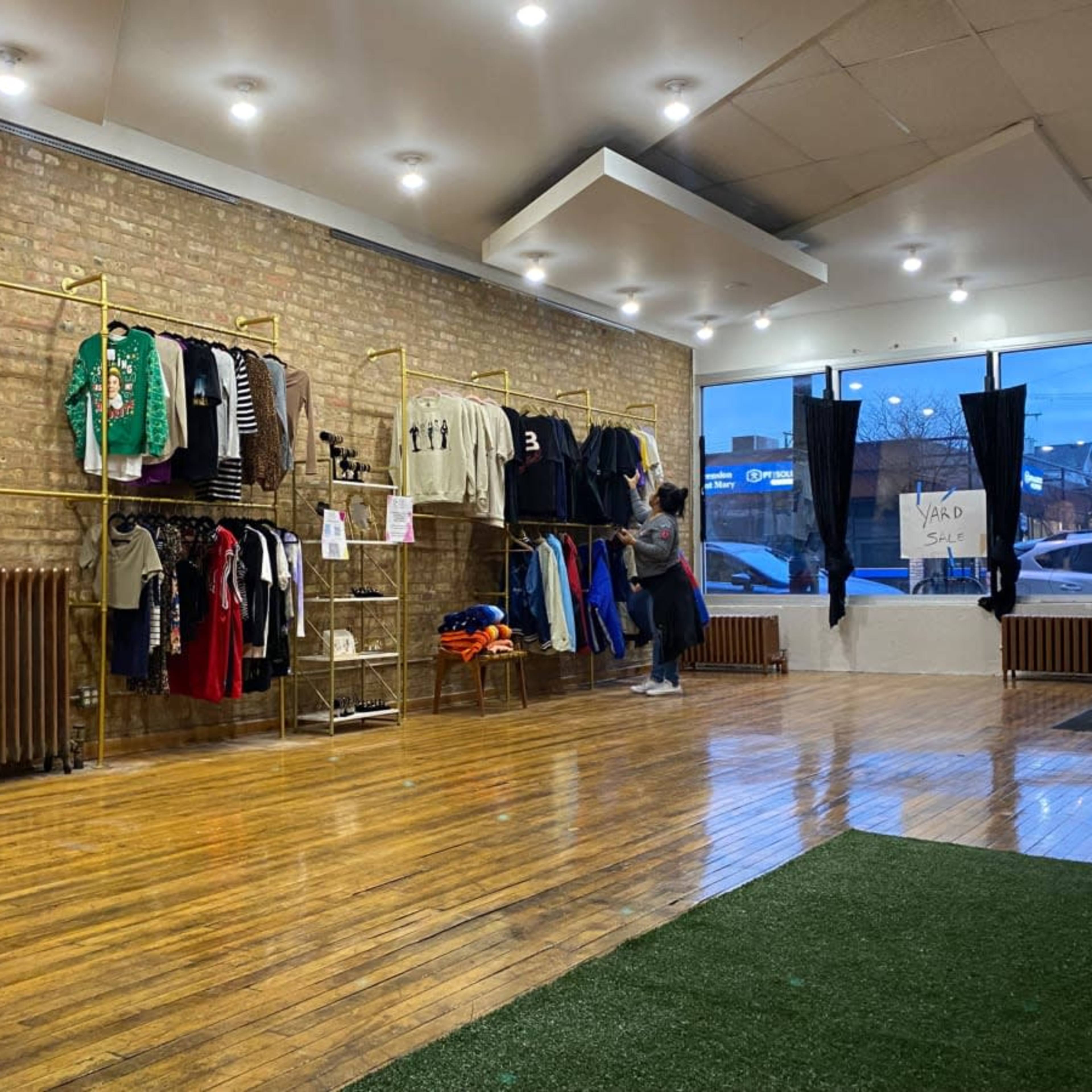 An interior of a clothing store with racks displaying various apparel along a brick wall, and large windows that provide natural light.
