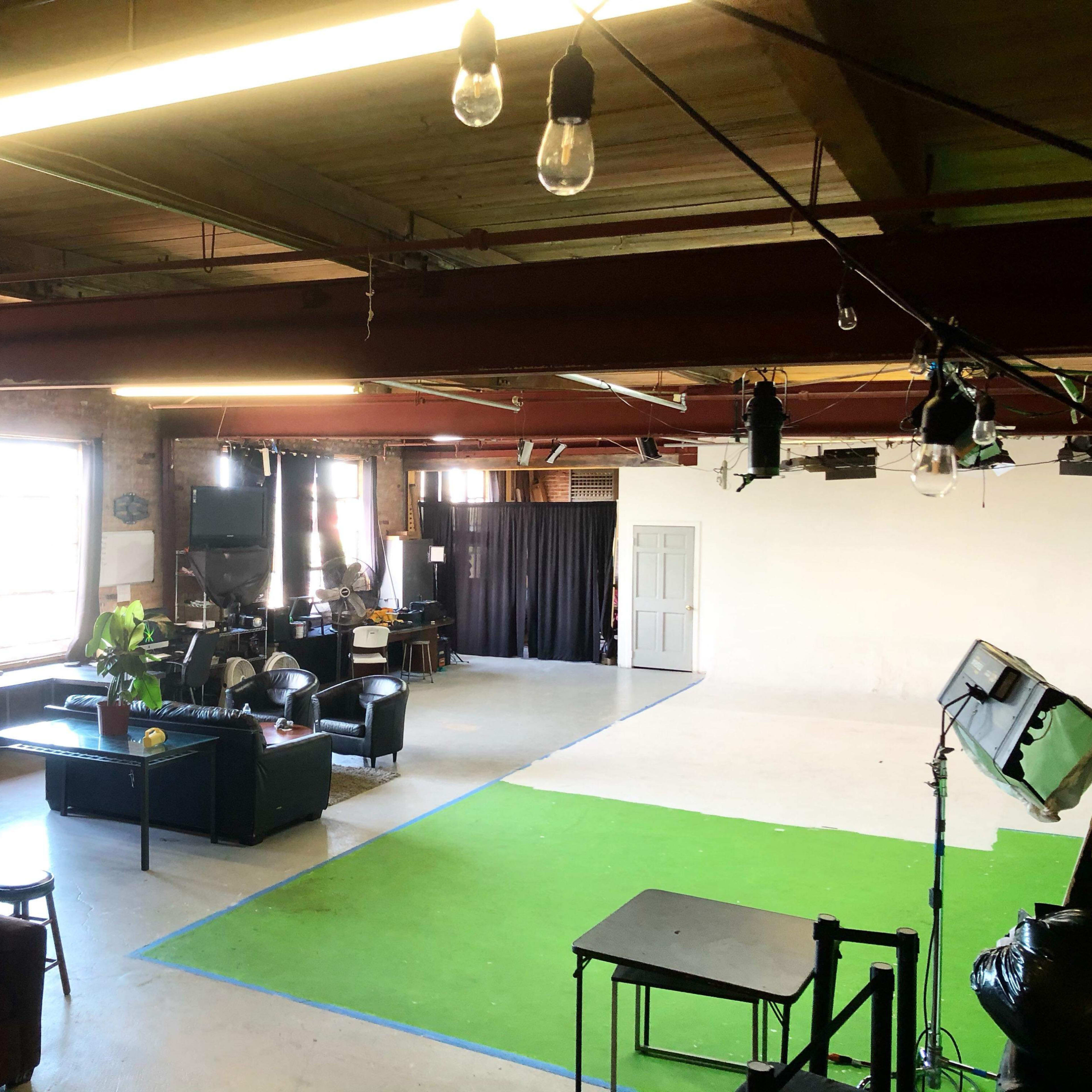 A spacious studio with a green carpet area, a black couch, and lighting equipment set up for filming.