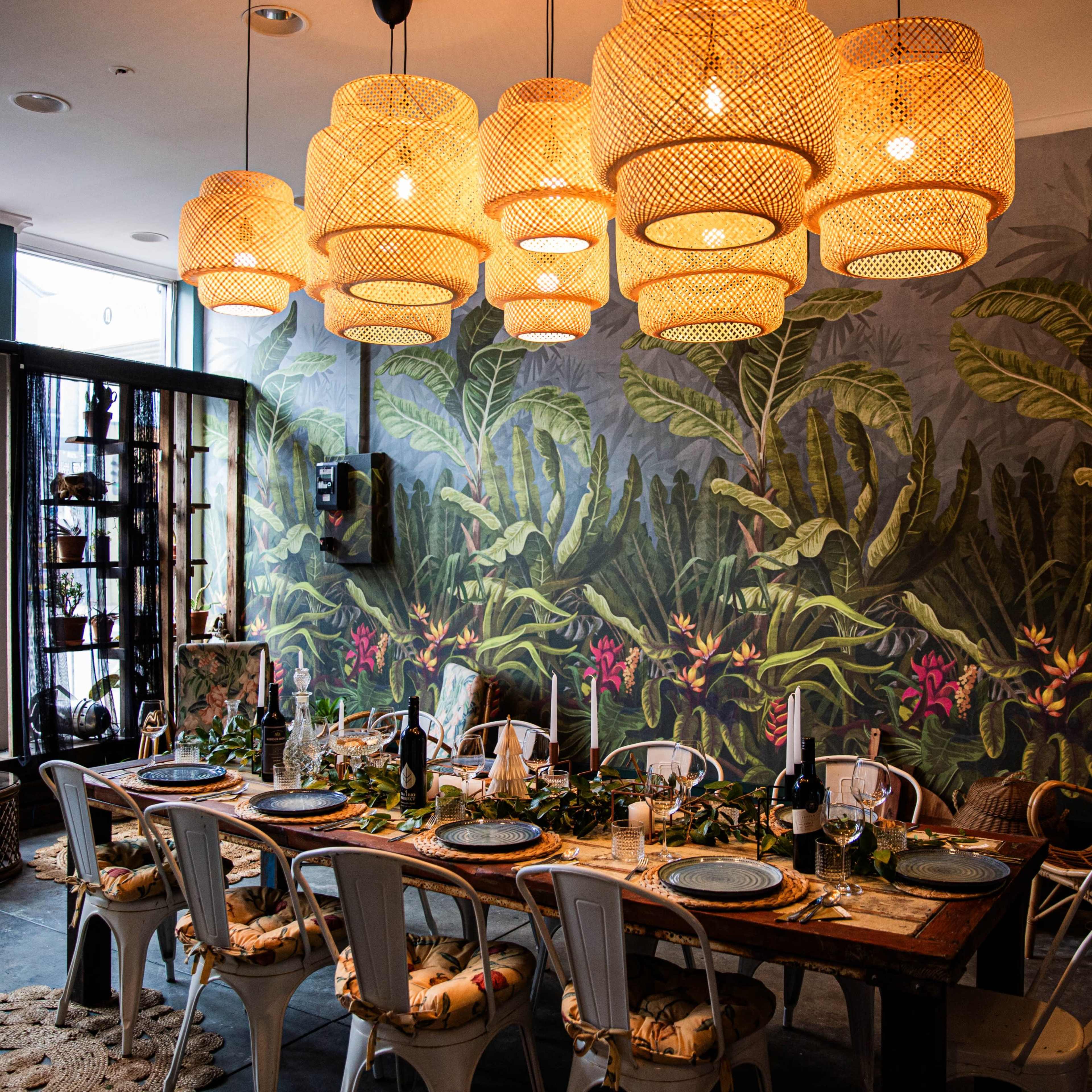 A dining area features a long wooden table set for a meal, surrounded by white chairs, with a vibrant tropical mural and large woven pendant lights overhead.