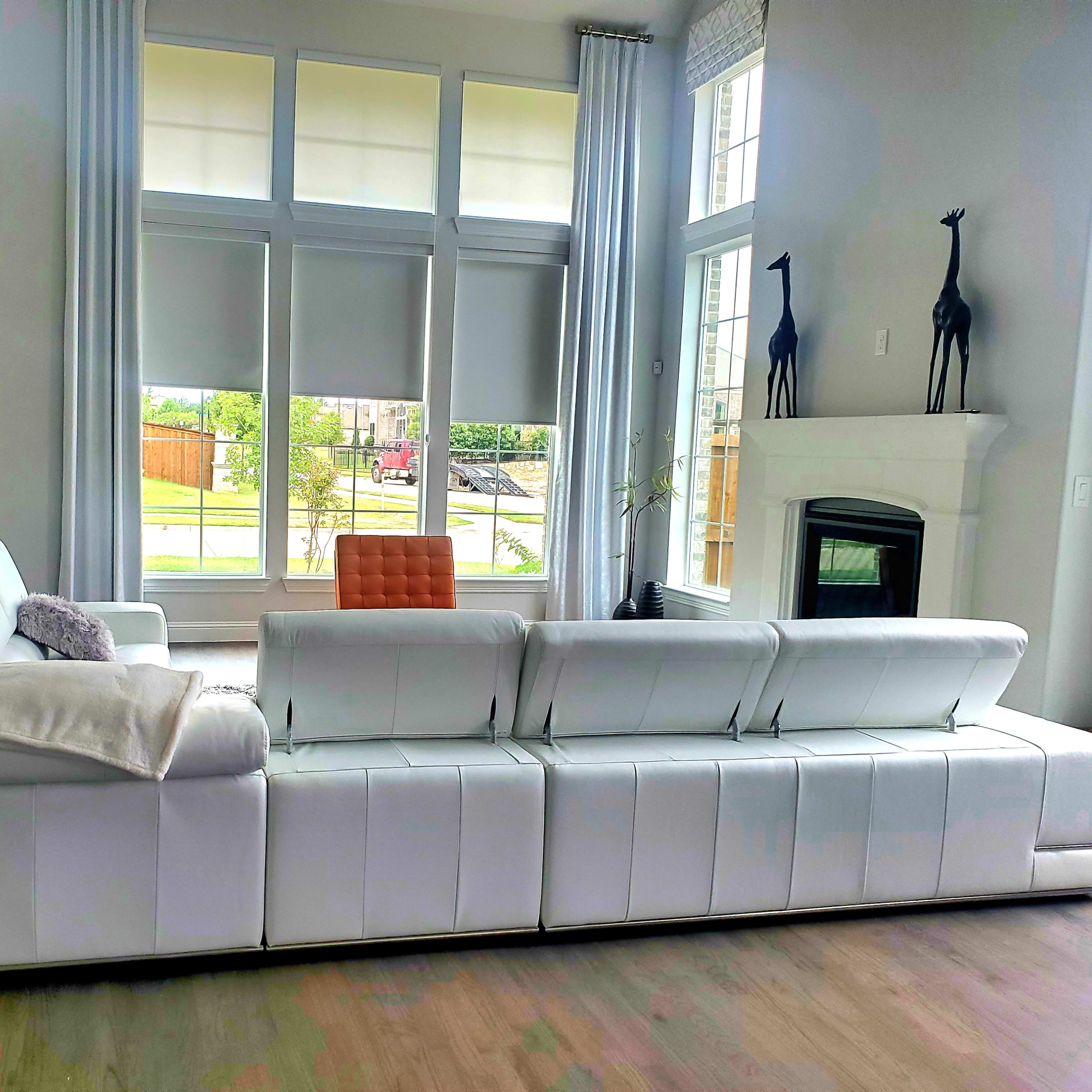 A modern living room features a spacious white sectional sofa facing a large window with vertical blinds and a white fireplace.