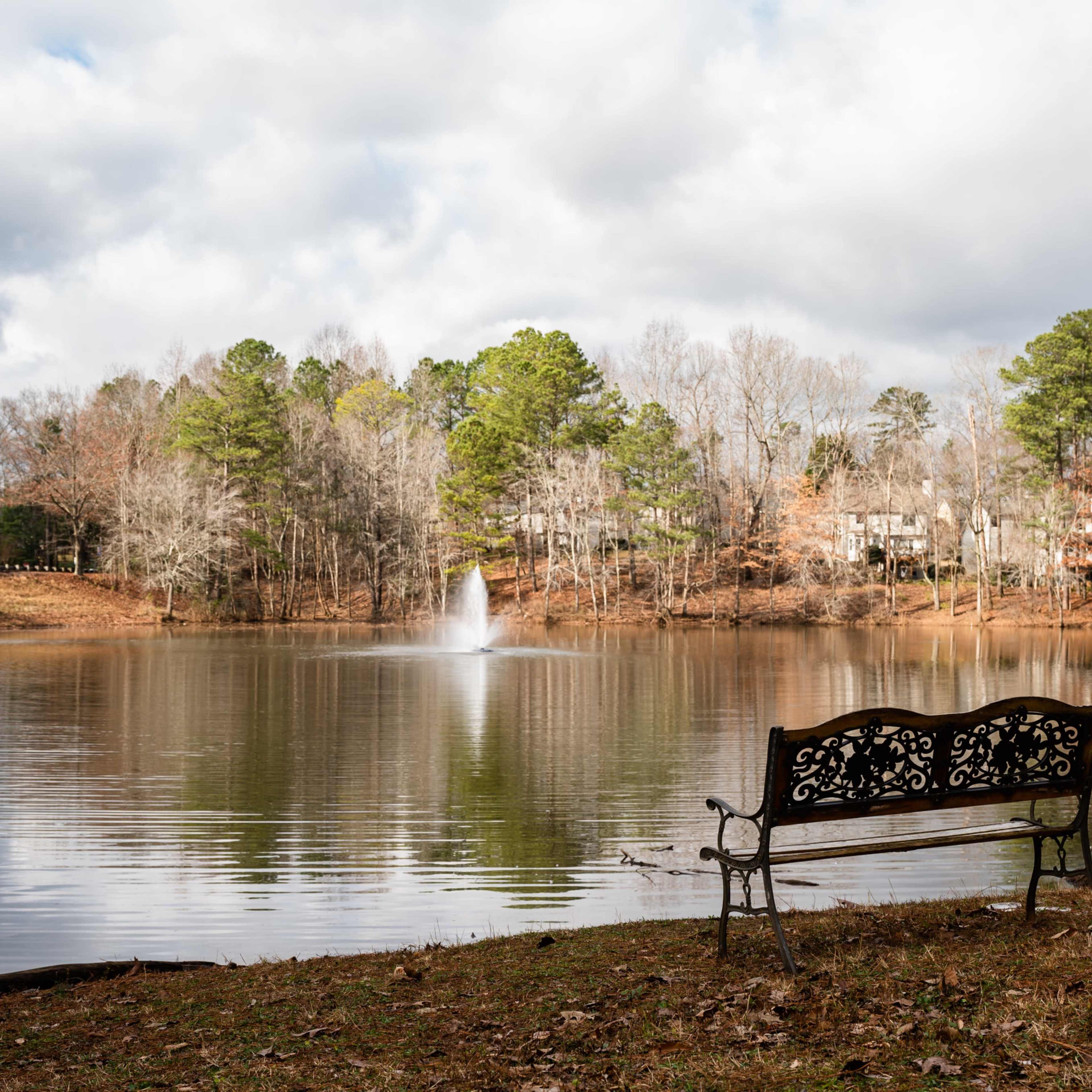 A wrought iron bench sits empty by a calm lake, with a fountain spraying water in the background and trees lining the shore.