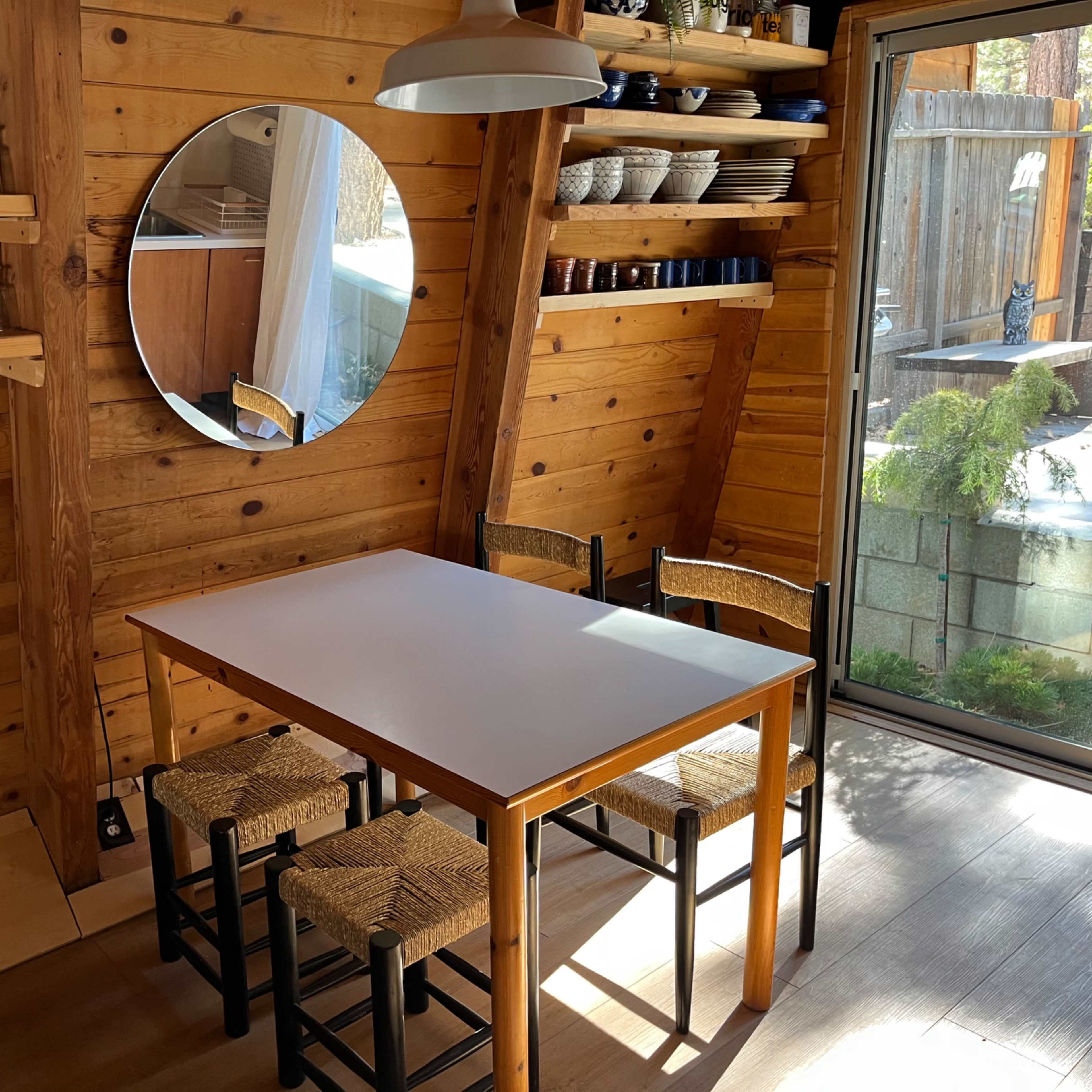 A simple dining area features a white table surrounded by four chairs in a wooden room with shelves holding glassware and dishes, and a large round mirror on the wall.