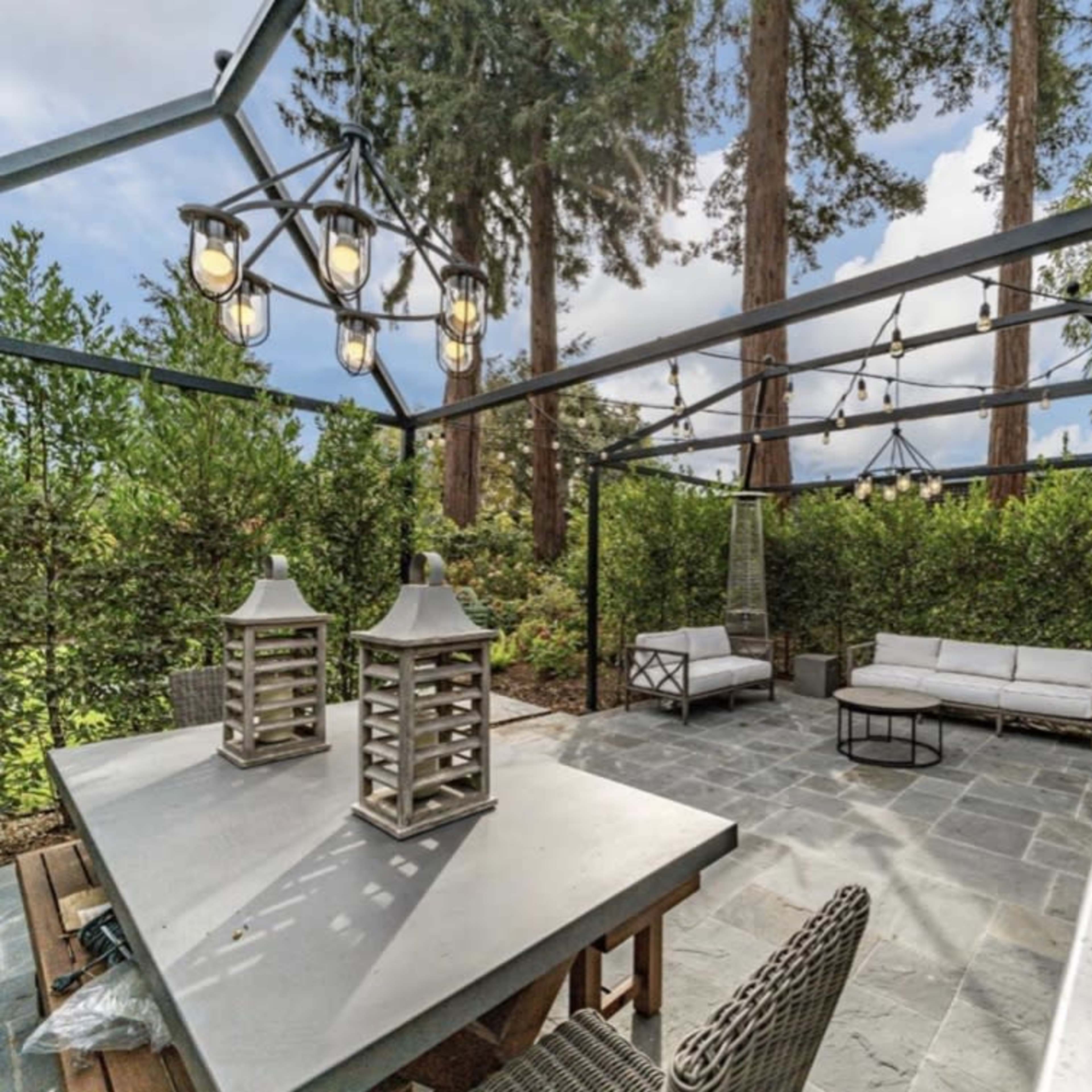 A covered outdoor patio area with a stone floor, modern seating, a barbecue grill, and decorative lanterns surrounded by tall trees and greenery.