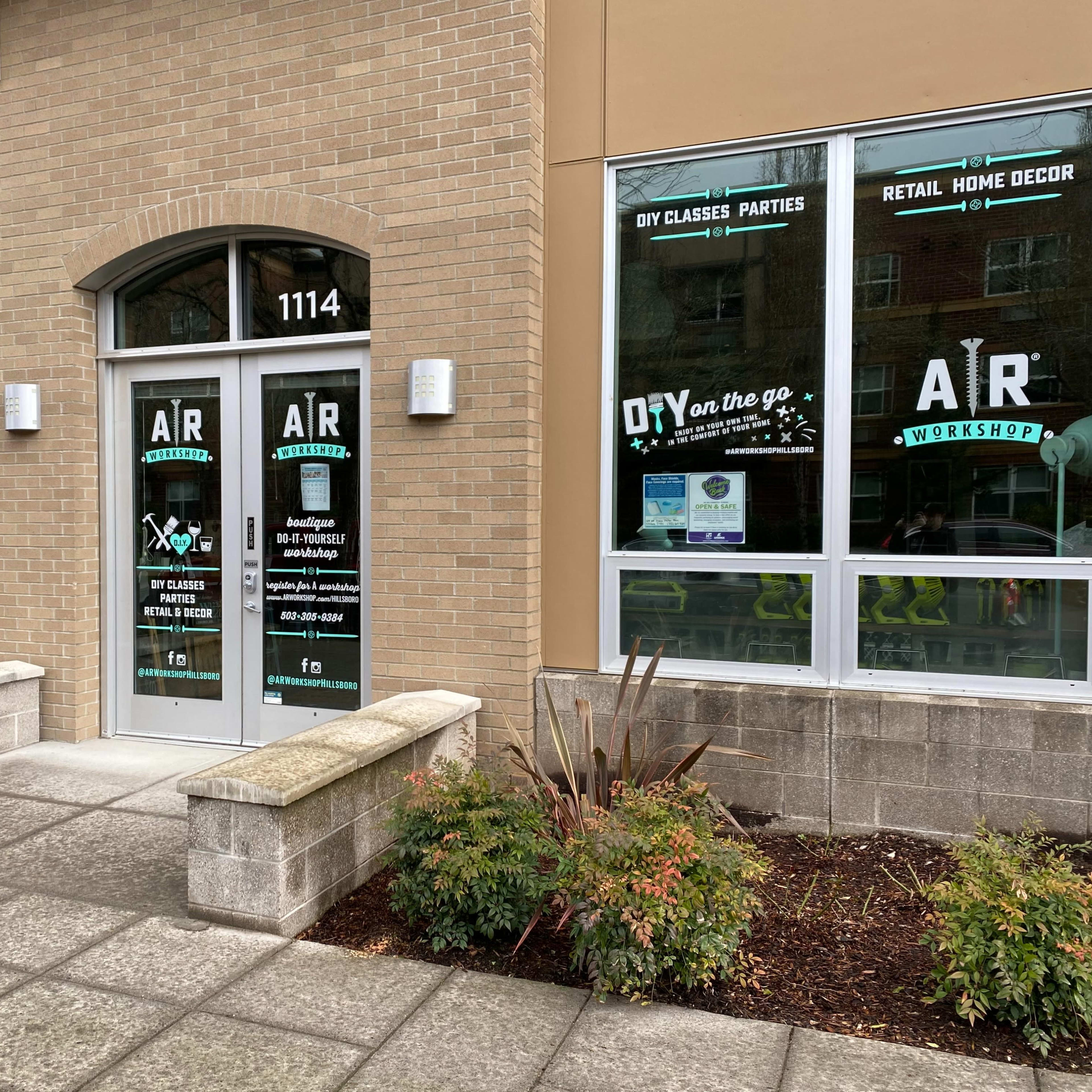 A storefront with large glass doors and windows displaying the business name "AR Workshop" along with promotional graphics for DIY classes and home decor.