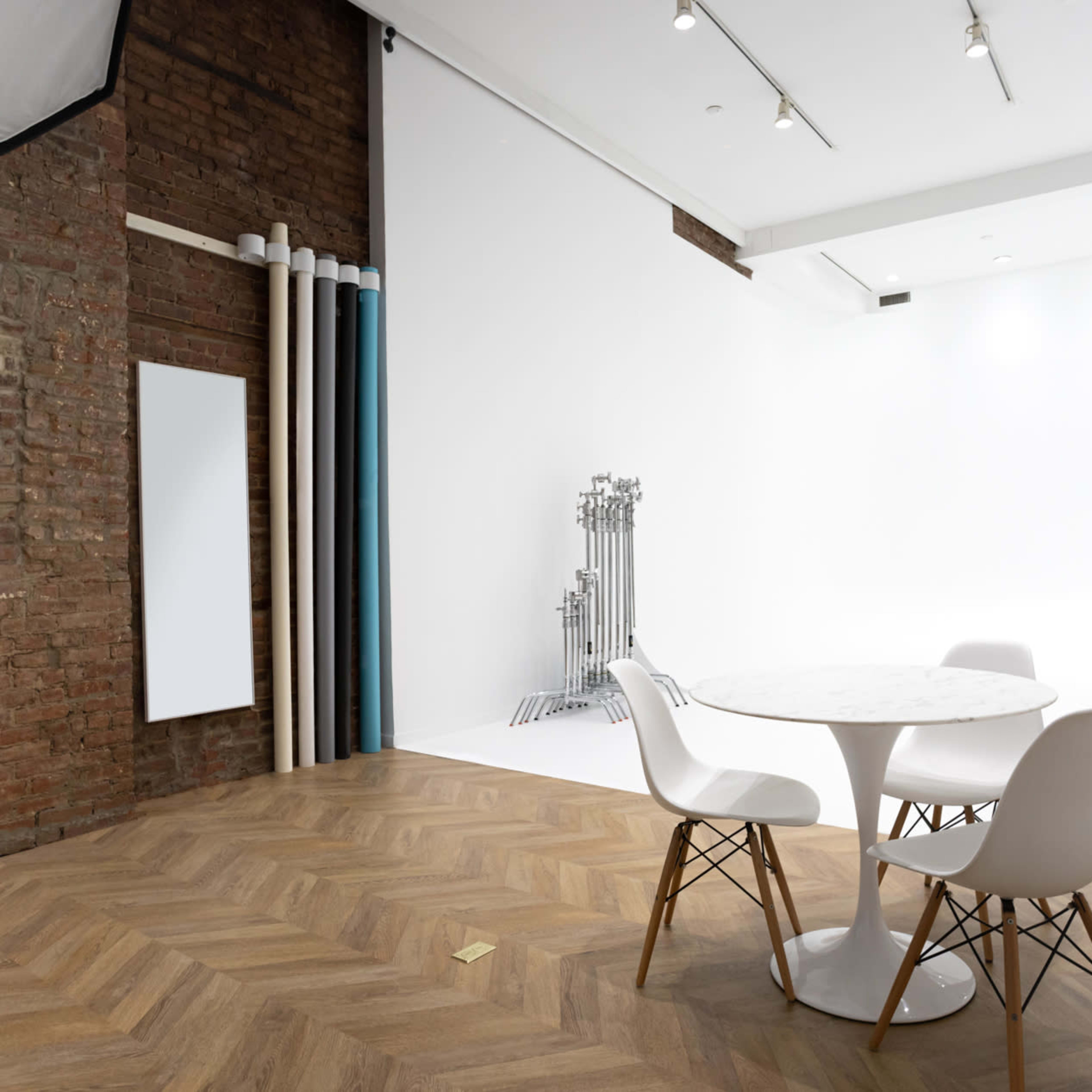 A modern photography studio with a round table and white chairs, featuring a brick wall and a large white backdrop.