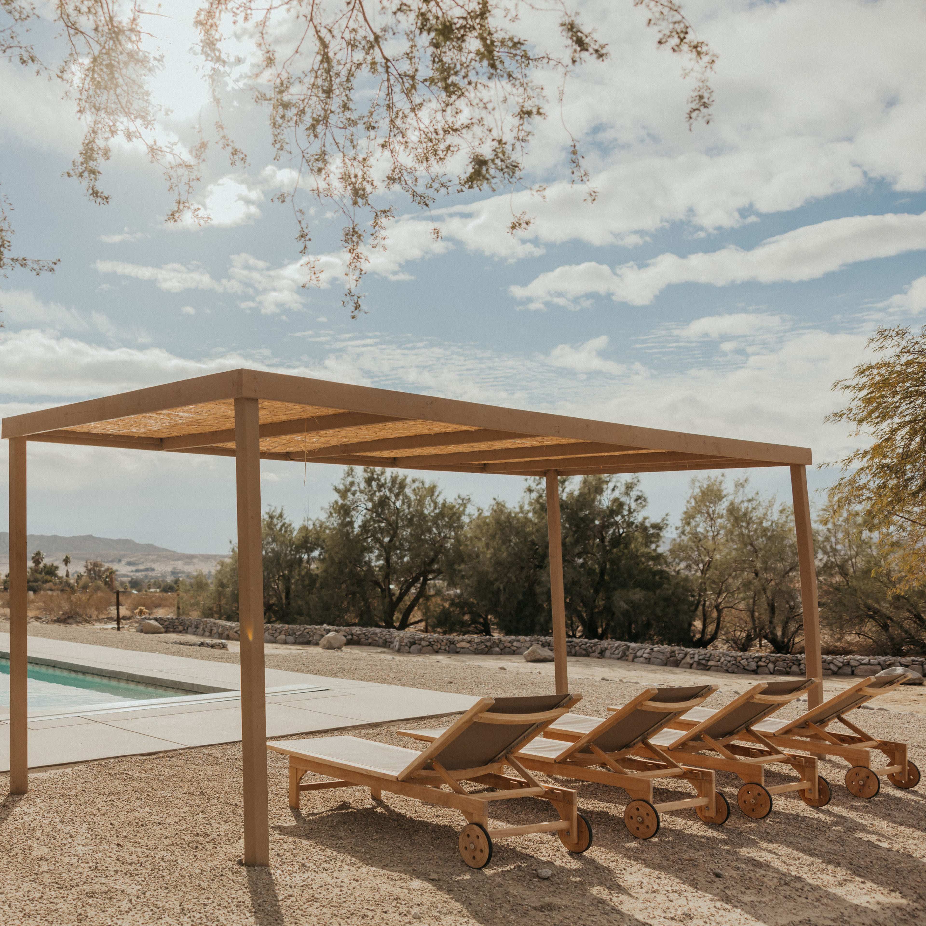 A simple wooden pergola shades a row of lounge chairs next to a rectangular swimming pool surrounded by a gravel landscape.