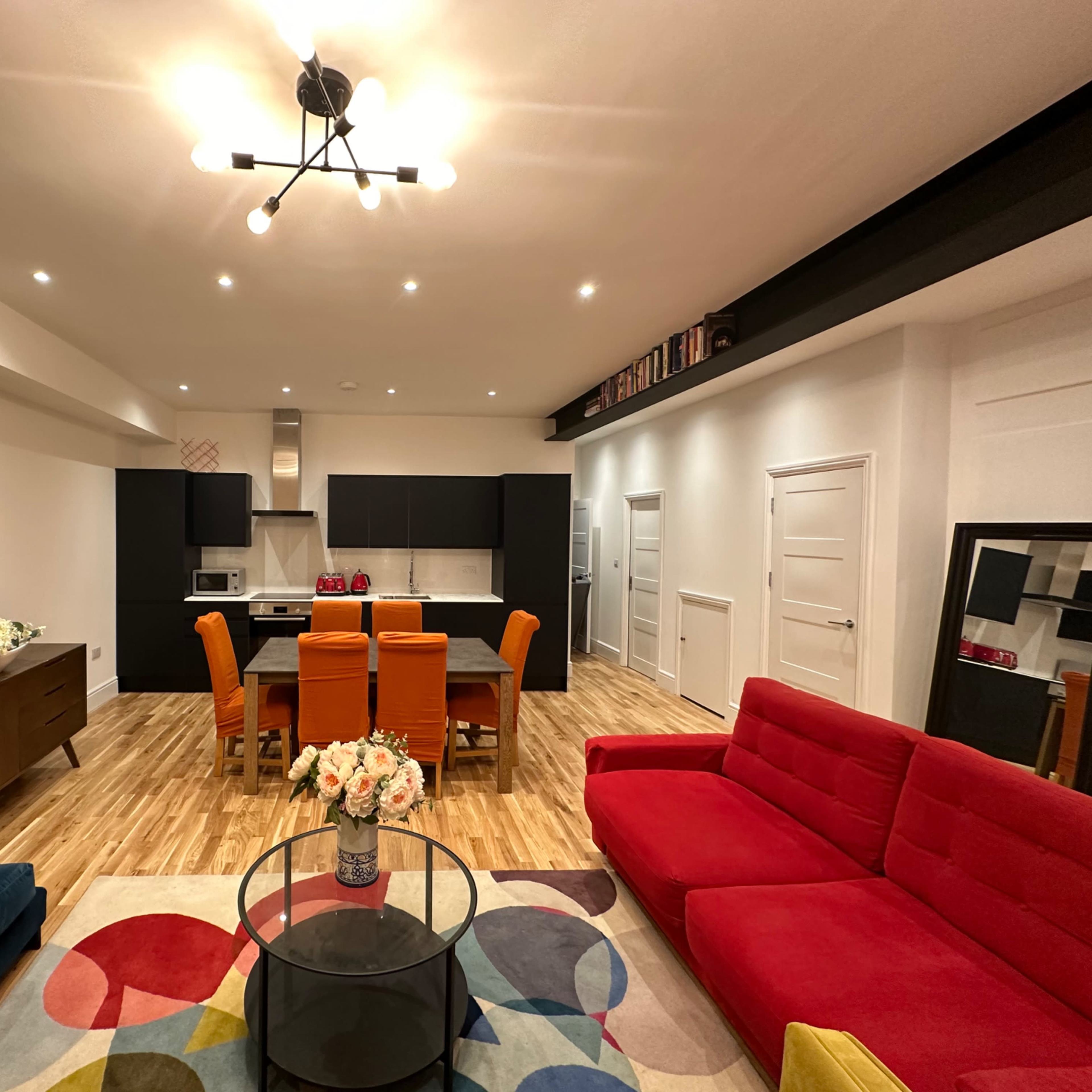 A modern living space featuring a red sofa, a blue couch, a circular coffee table, a dining area with orange chairs, and a kitchen in the background.