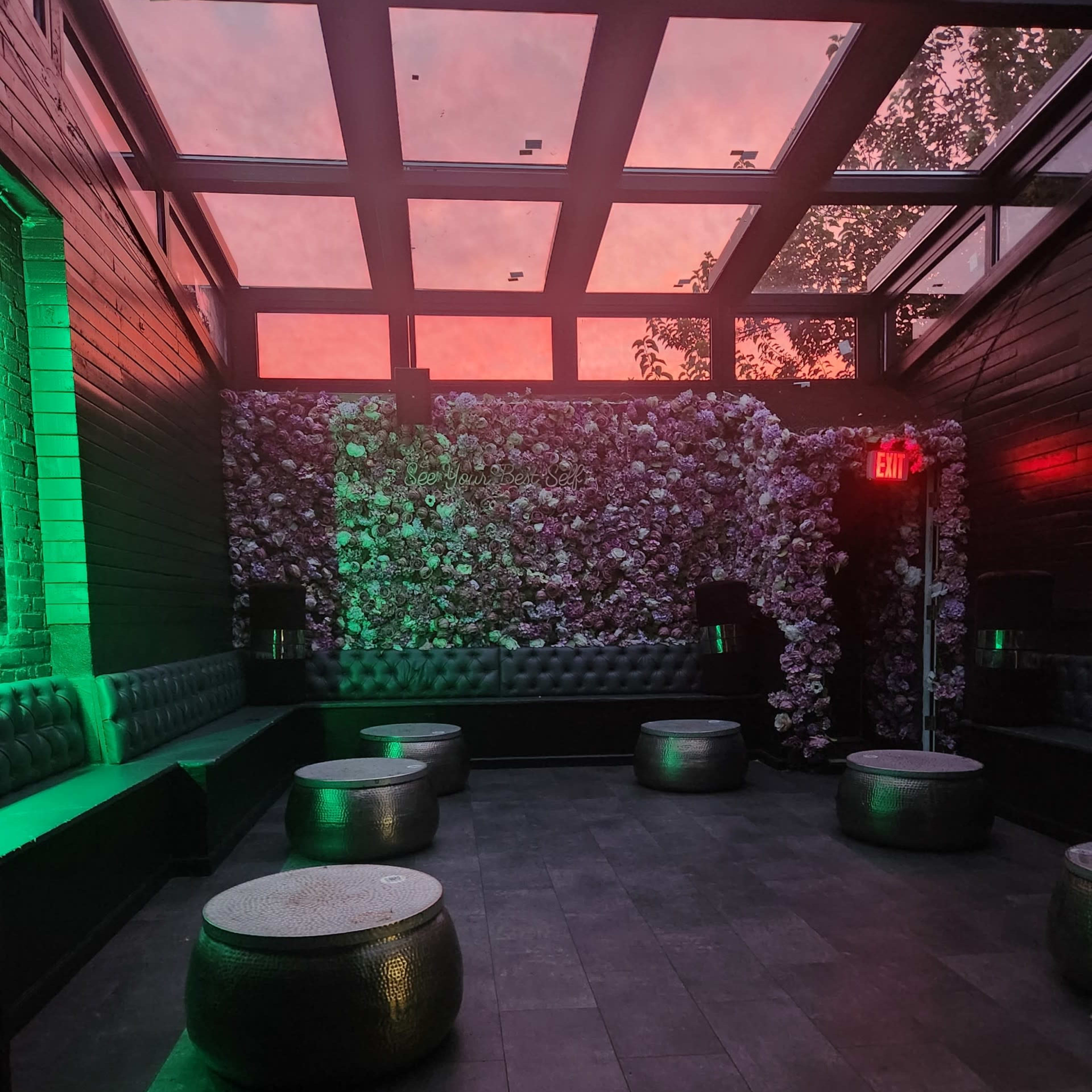 A dimly lit lounge area with floral wall decor, circular tables, and a glass ceiling reflecting a pinkish sky.
