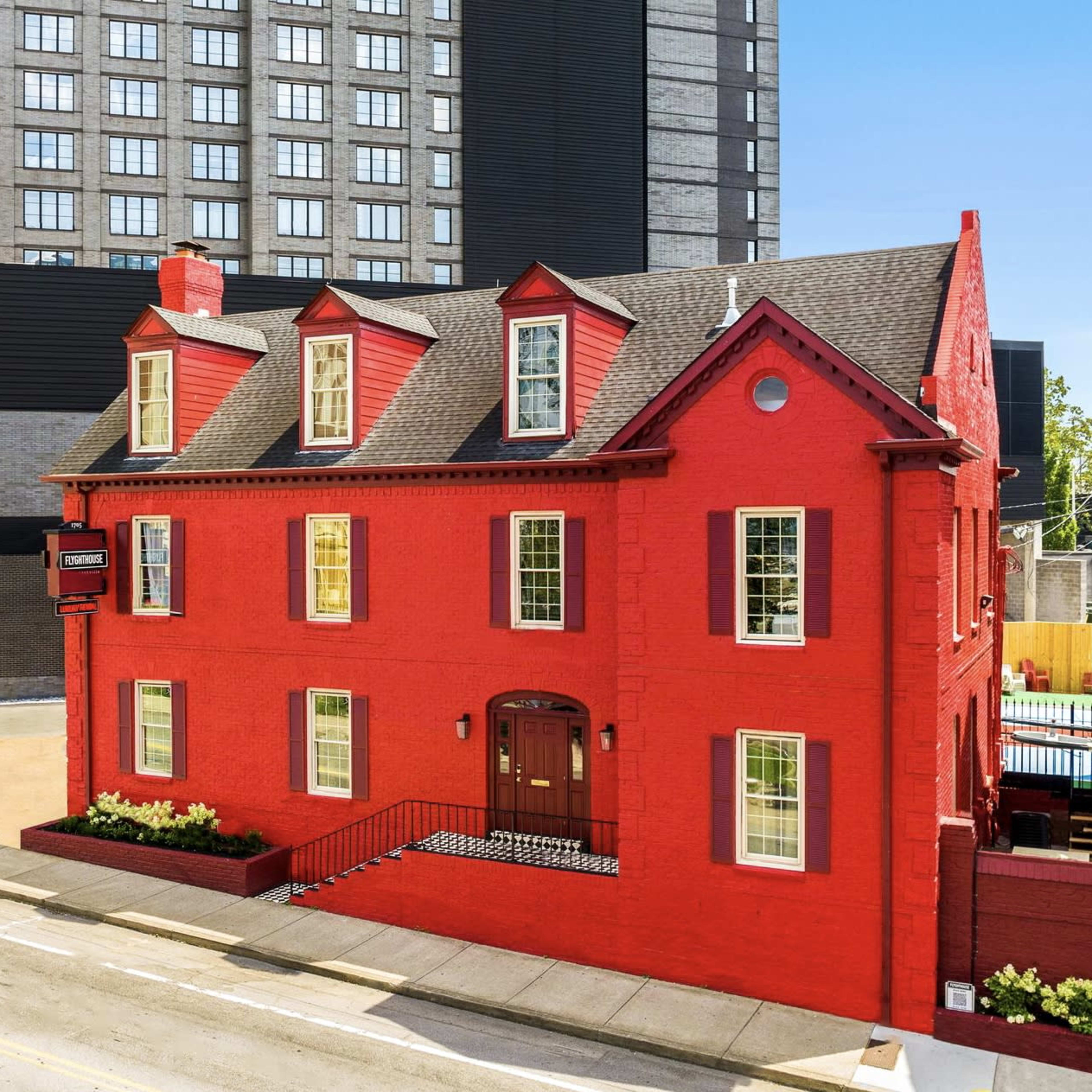 A red brick house with a steep roof and multiple windows, situated next to a tall, modern building.