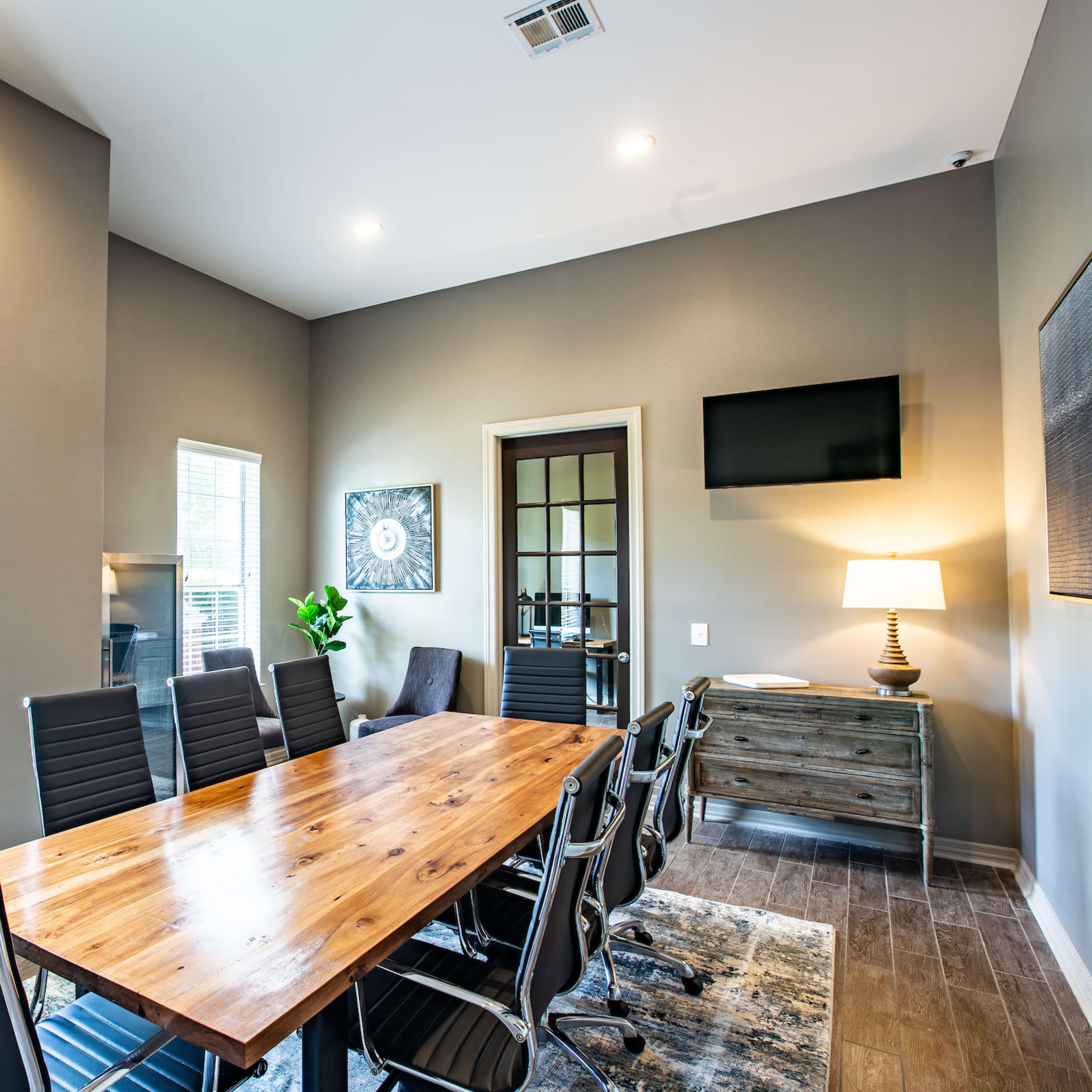 A modern conference room features a long wooden table surrounded by black chairs, with large windows and a TV mounted on the wall.