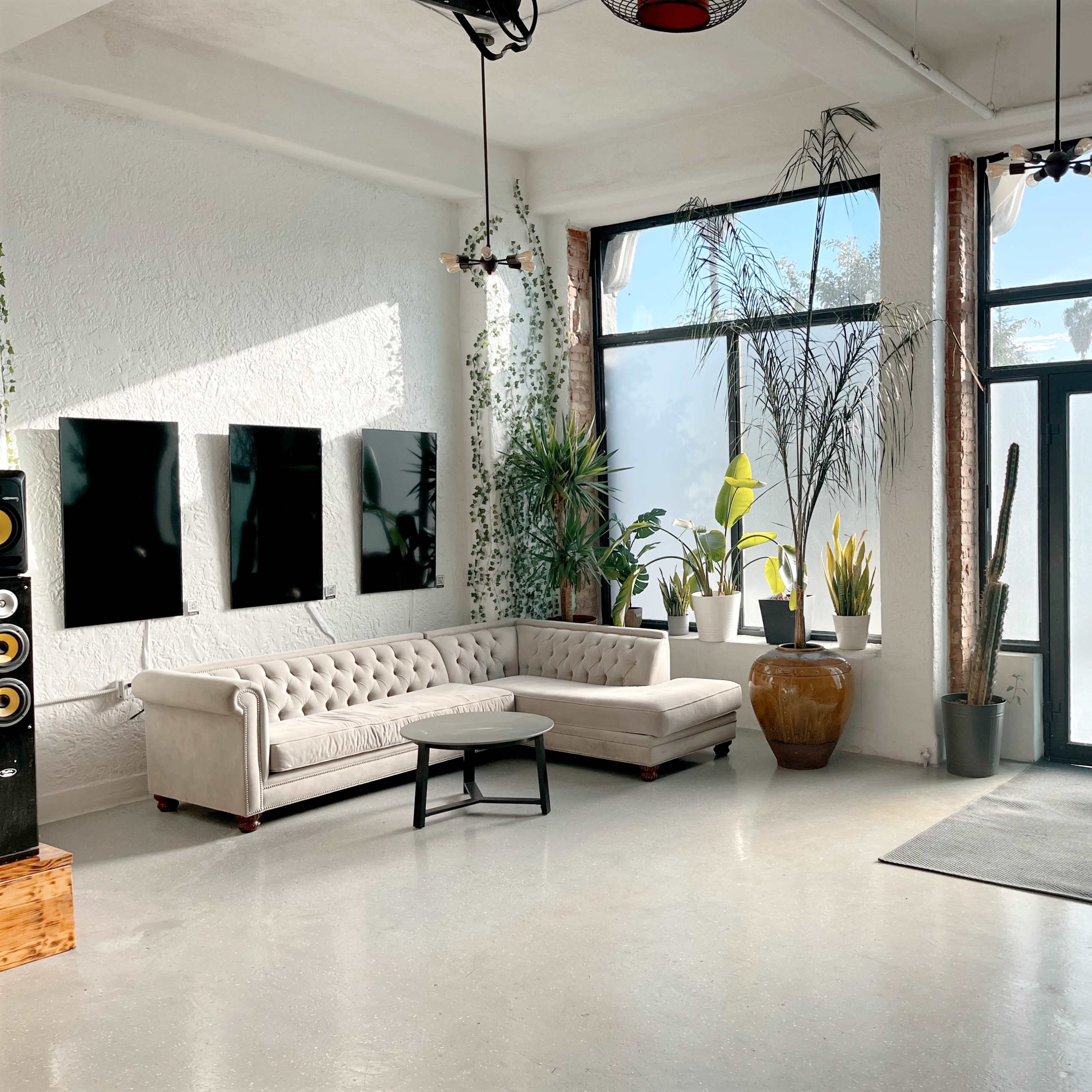 A bright, modern interior with a light-colored sofa, a round table, and large windows surrounded by various plants.