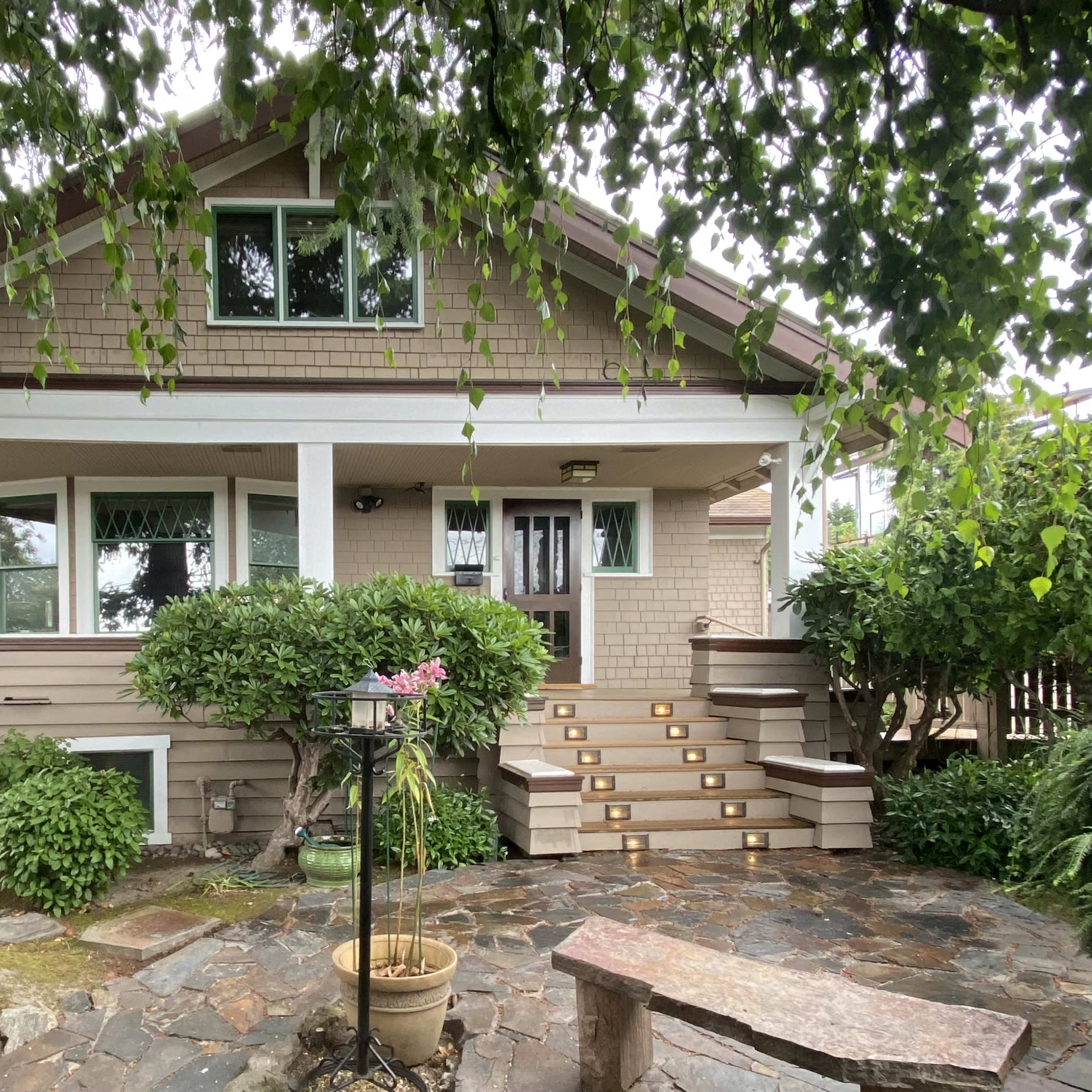 A two-story house with a front porch, surrounded by lush greenery and stone pathways.
