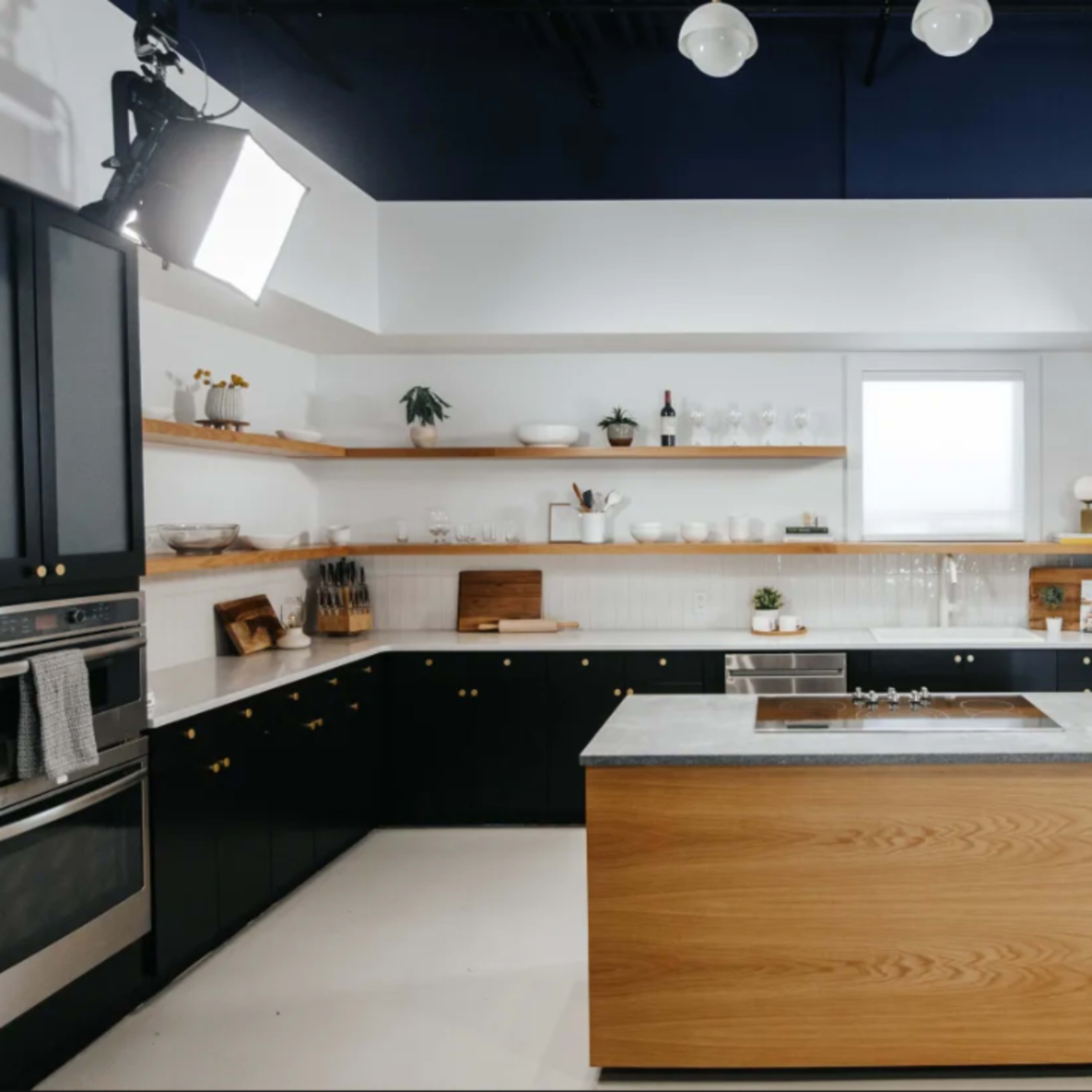 A modern kitchen featuring black cabinetry, a central wooden island, and stainless steel appliances, with shelves displaying dishware.