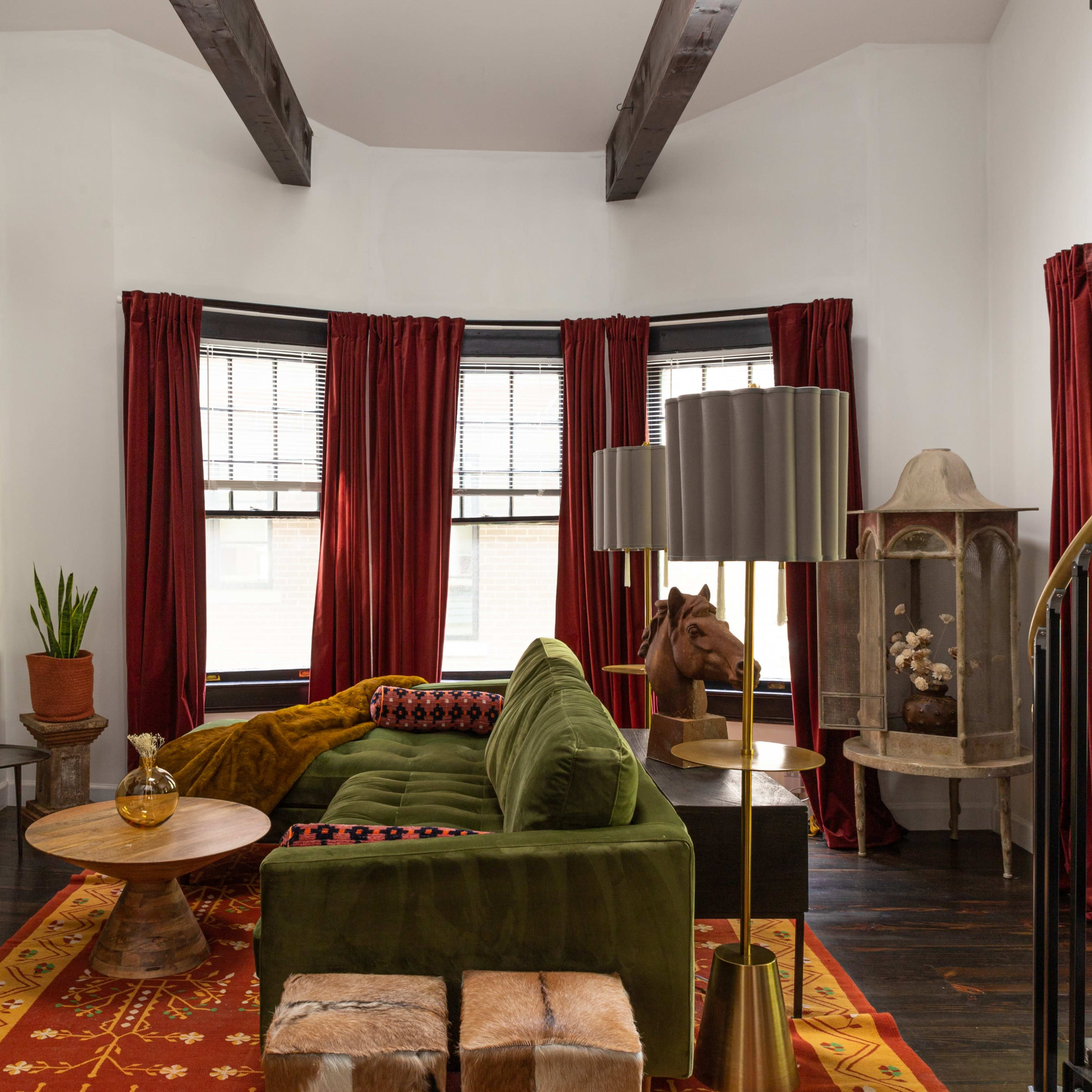 A living room with a green sofa, a round coffee table, and large windows draped with red curtains, featuring wooden beams on the ceiling.