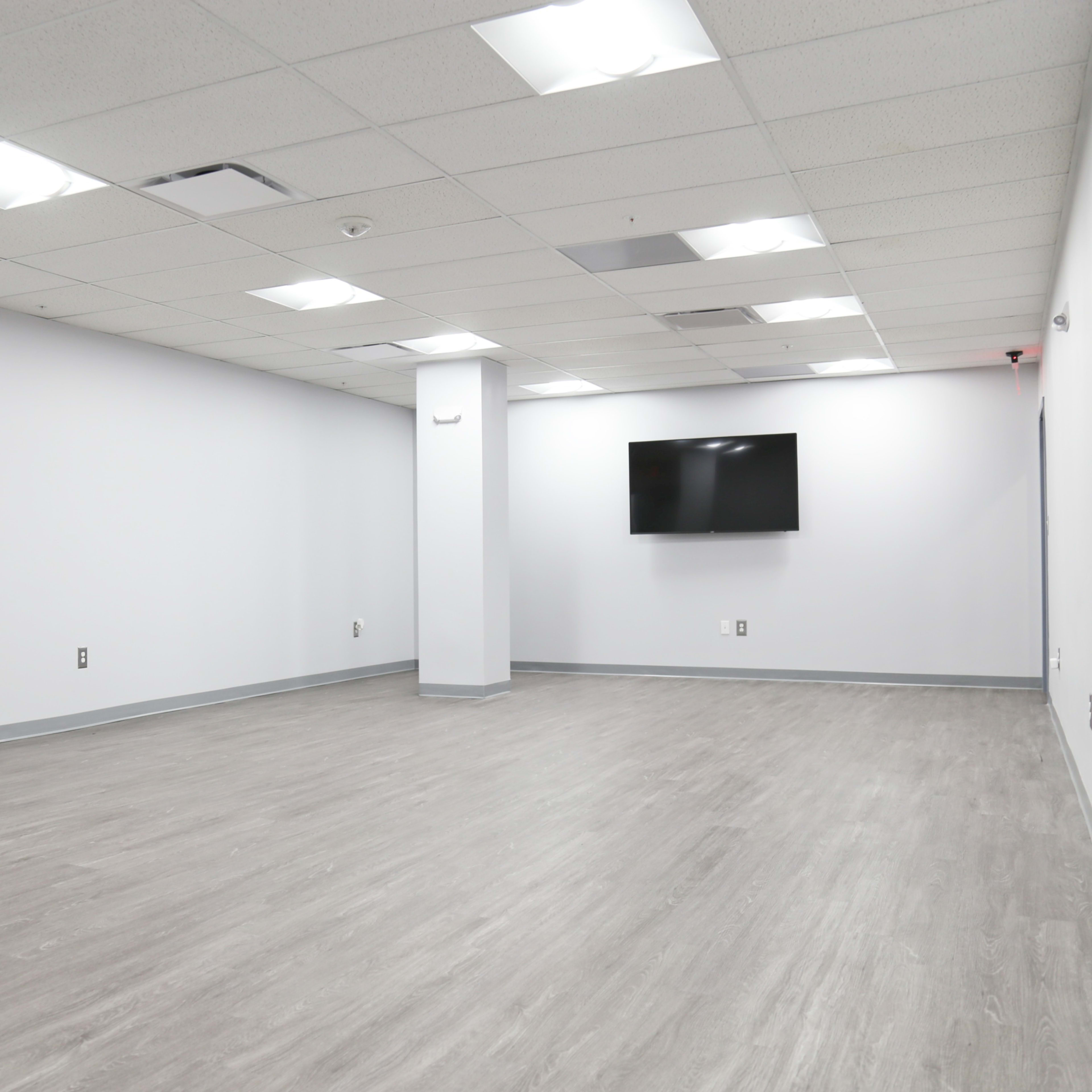 An empty, well-lit room with gray flooring, white walls, a wall-mounted television, and no furniture.