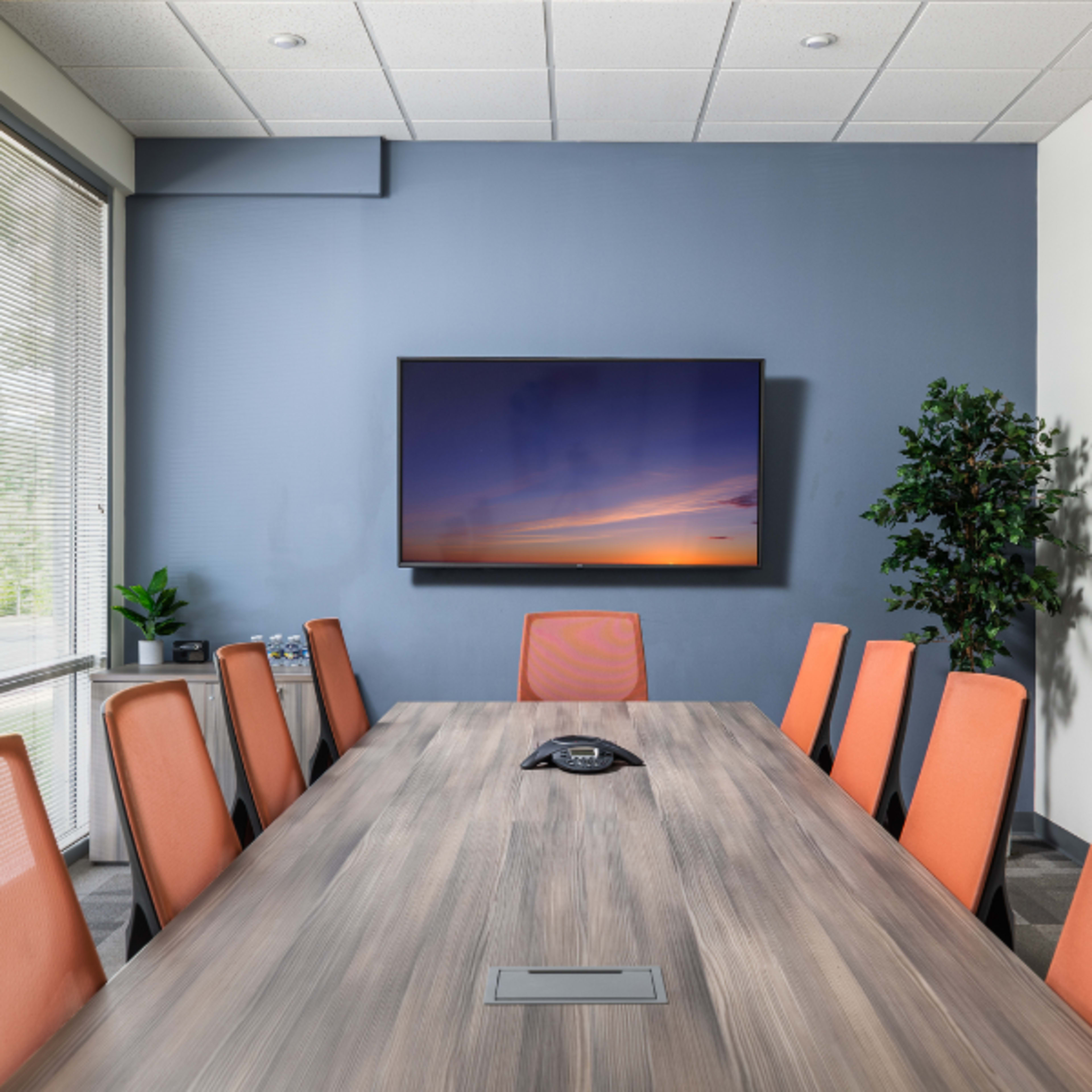A modern conference room features a long wooden table surrounded by orange chairs, with a large screen mounted on the wall and a whiteboard nearby.