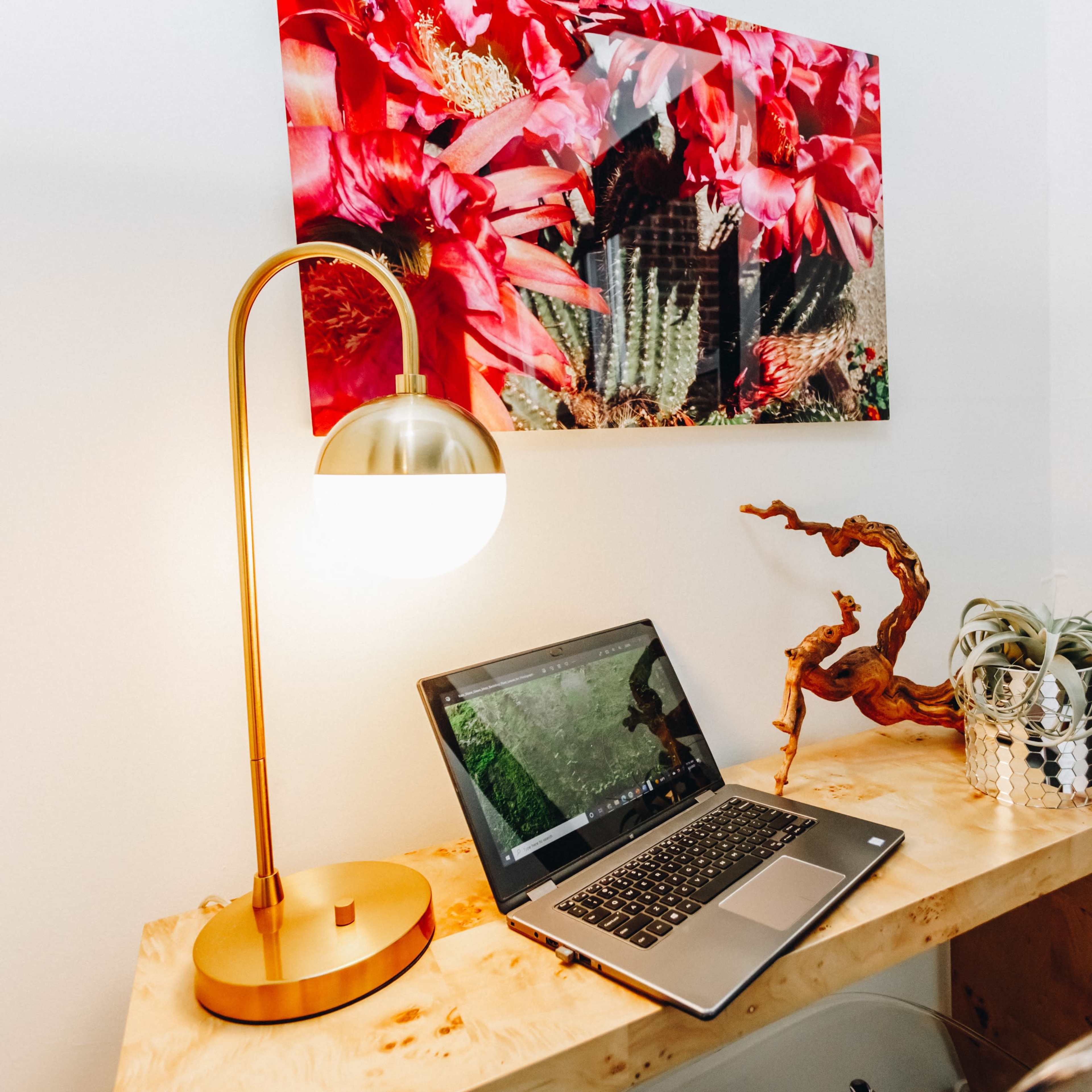 A wooden desk with a laptop, a decorative plant, and a lamp is positioned against a wall featuring a large floral artwork.