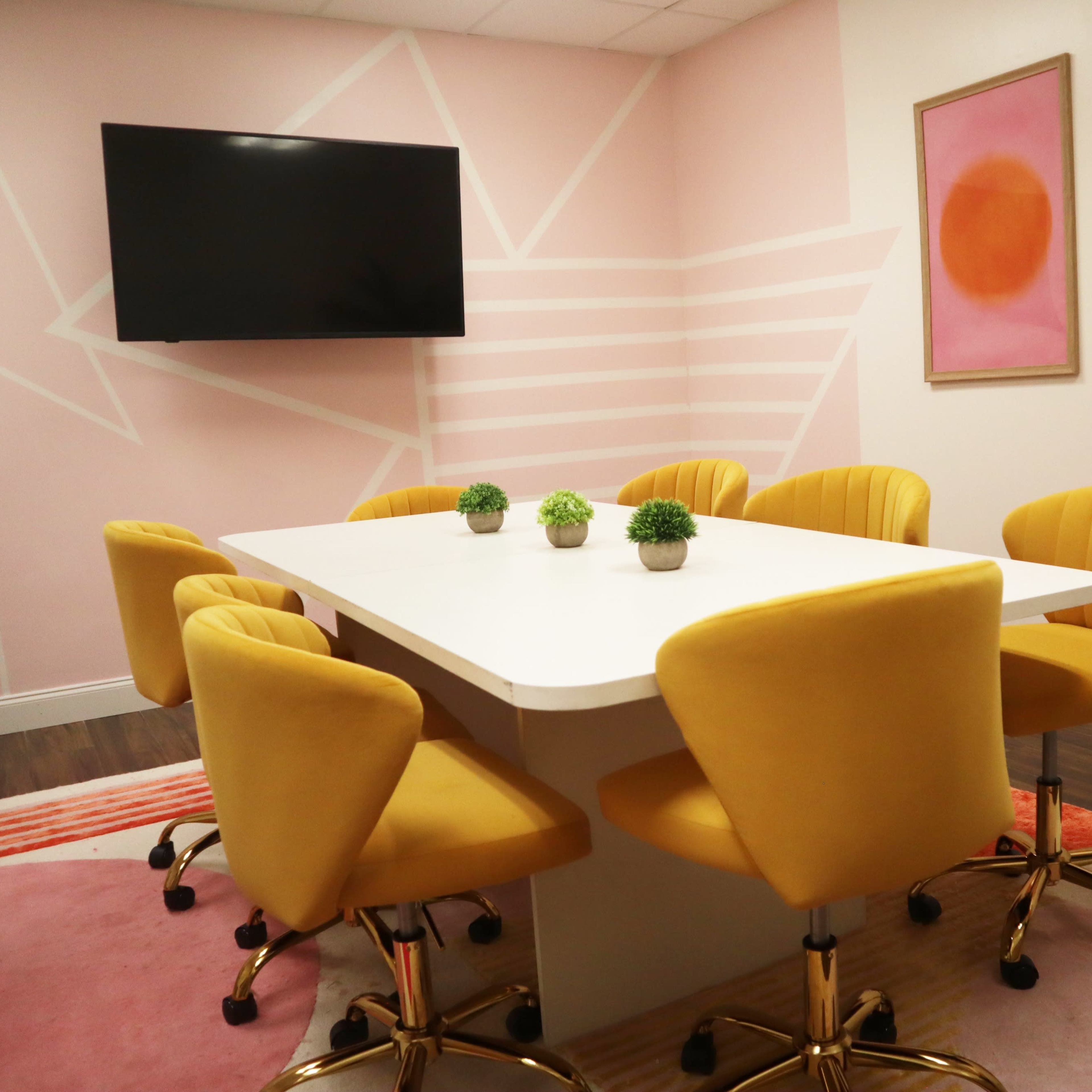 A modern conference room with a white table surrounded by eight yellow swivel chairs, a large wall-mounted TV, and colorful artwork.