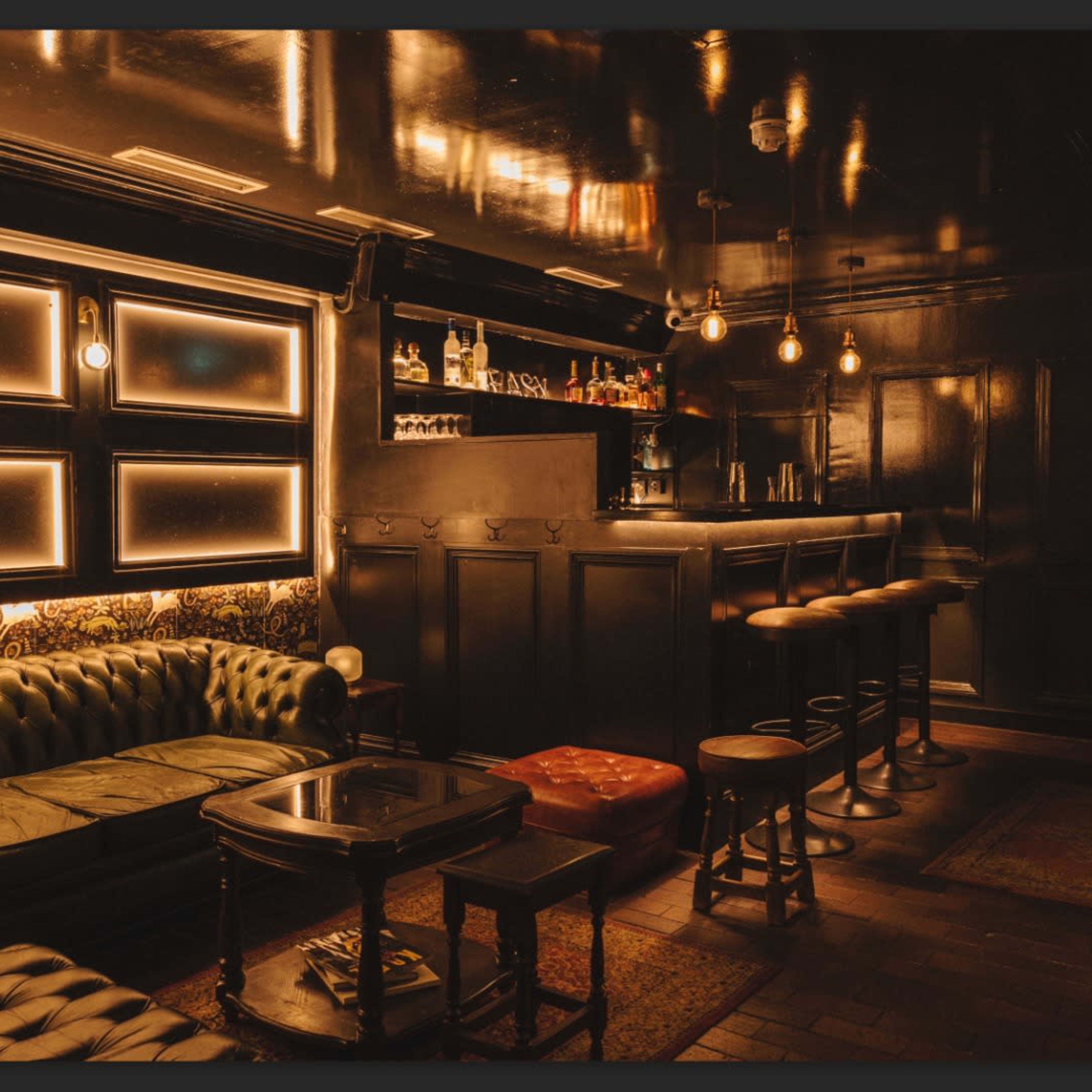 A dimly lit bar features a dark wood counter, a leather sofa, and several wooden stools arranged around a small table.