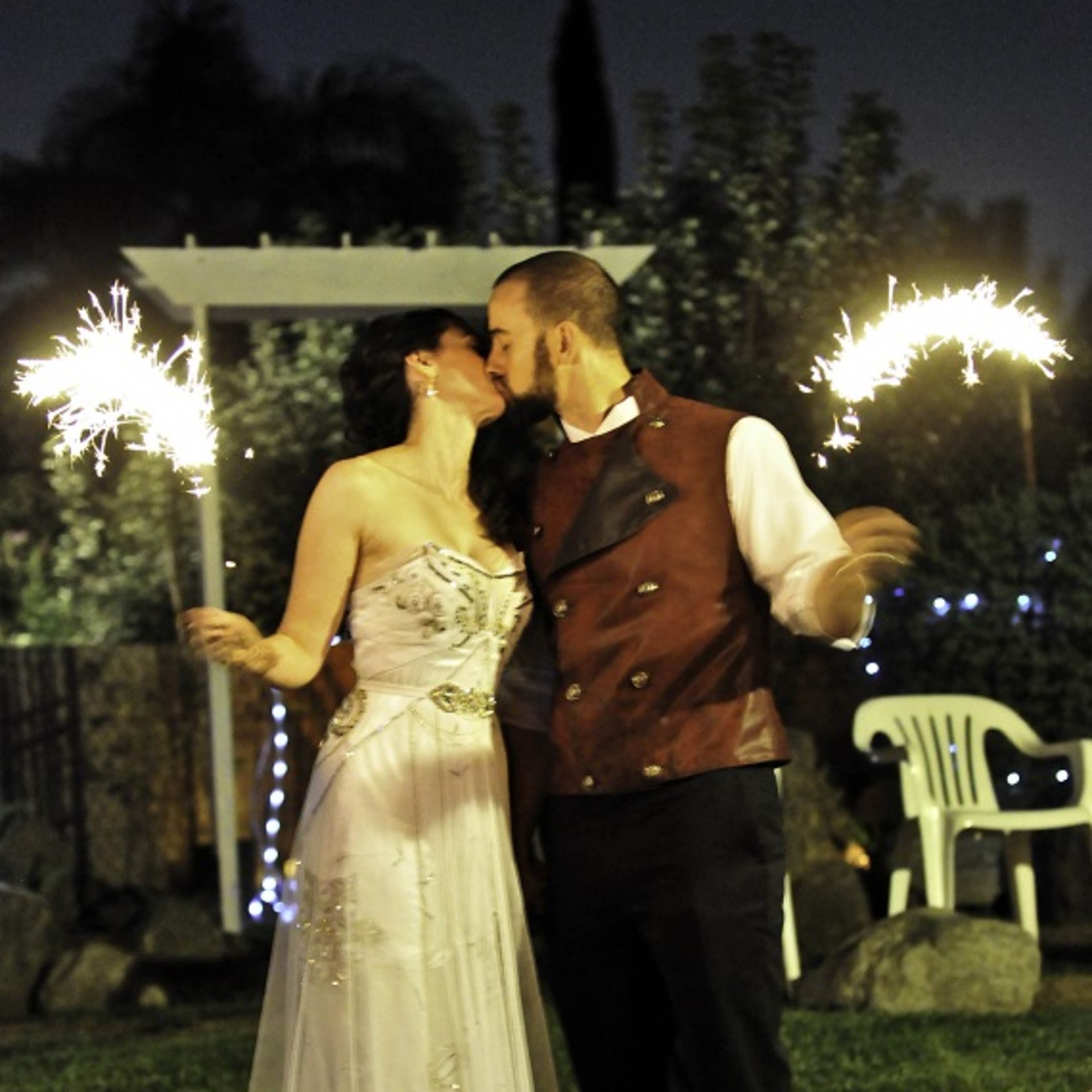 A couple in formal attire kisses while holding sparklers in a backyard illuminated by string lights.