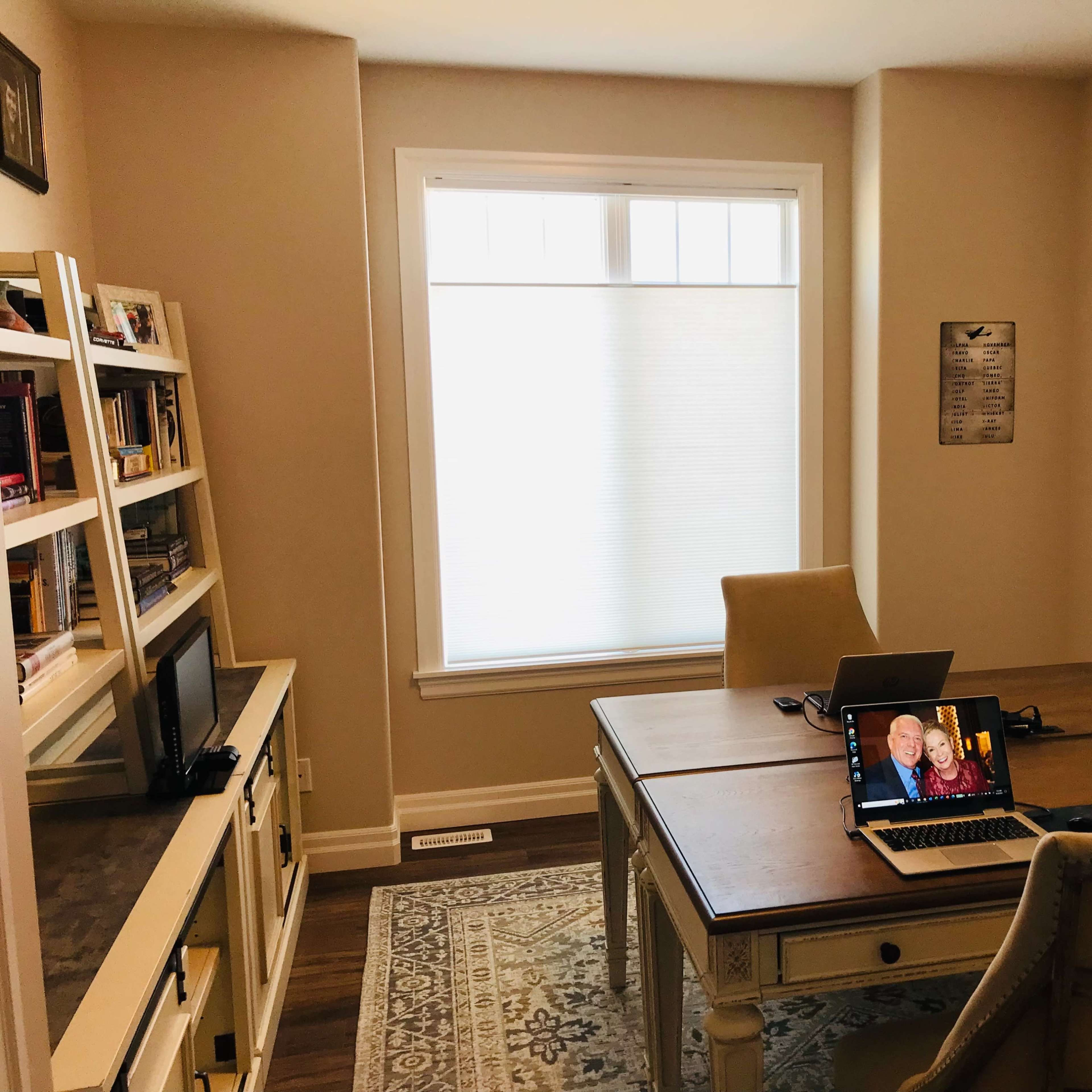 A small, light-colored office features a wooden desk with two laptops open, a bookshelf filled with books, and a window with shades letting in natural light.