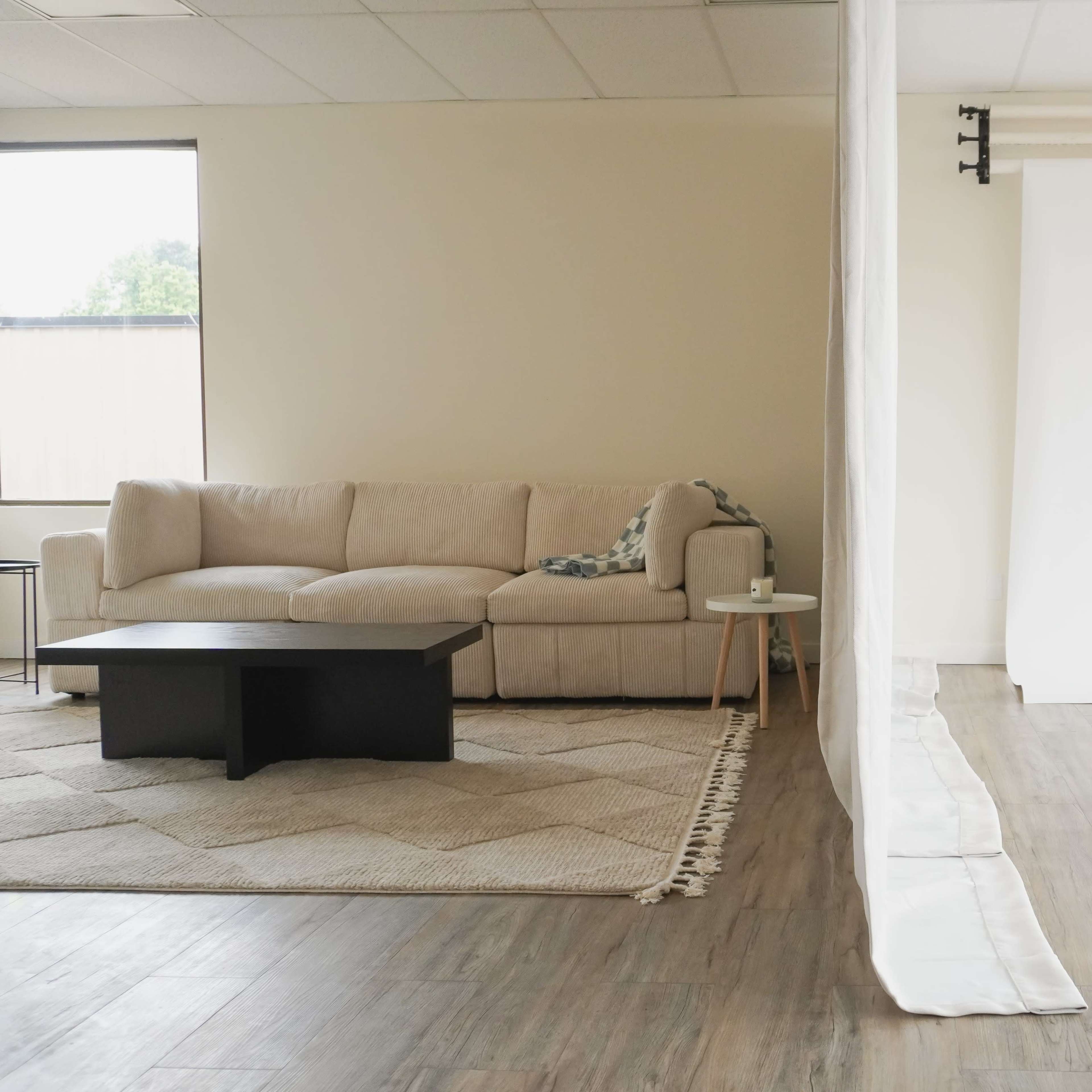 A spacious room divided by a white curtain, featuring a beige sofa, a black coffee table, and a white backdrop set up on one side.