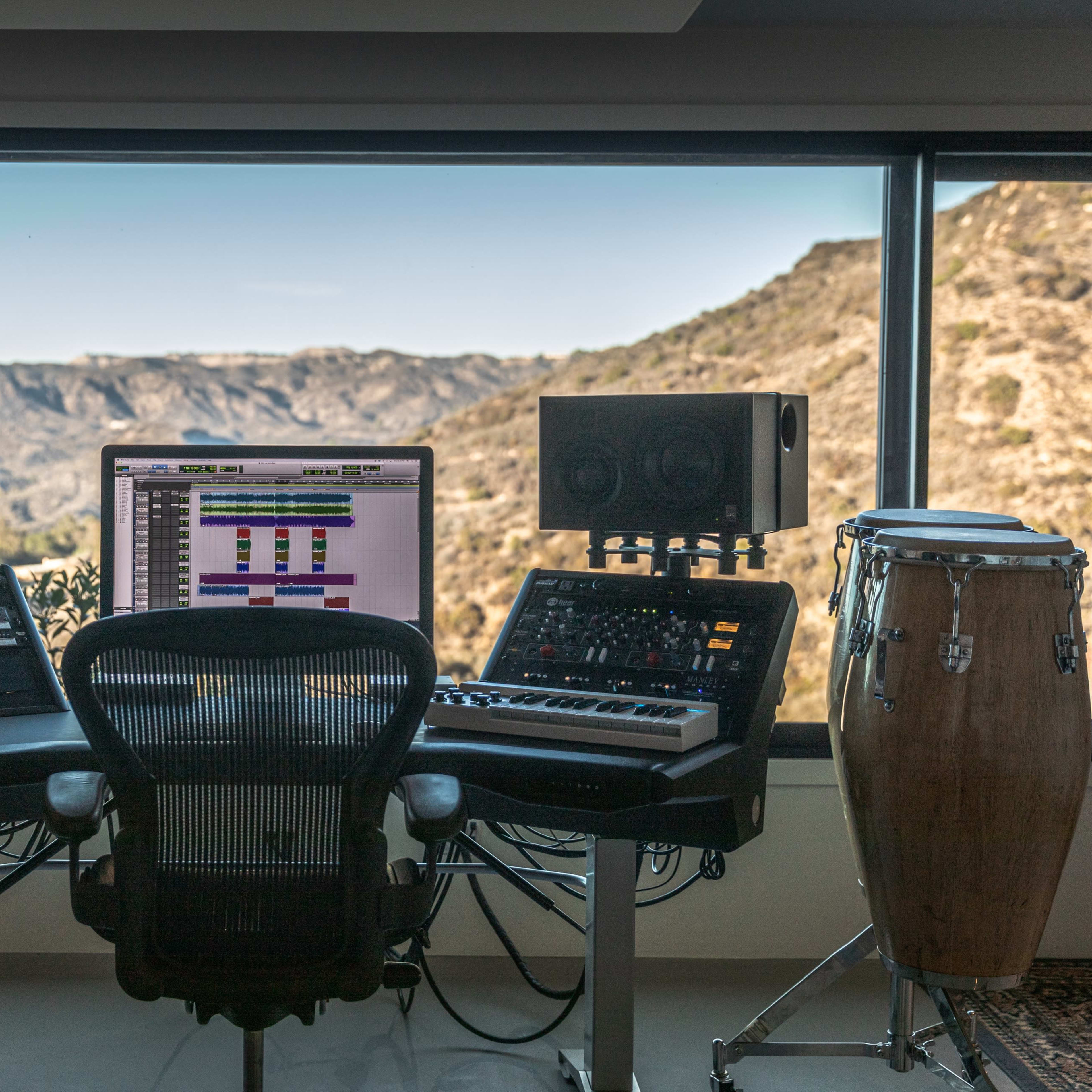 A modern music studio is set up with mixing equipment and instruments, facing large windows that showcase a mountain landscape.