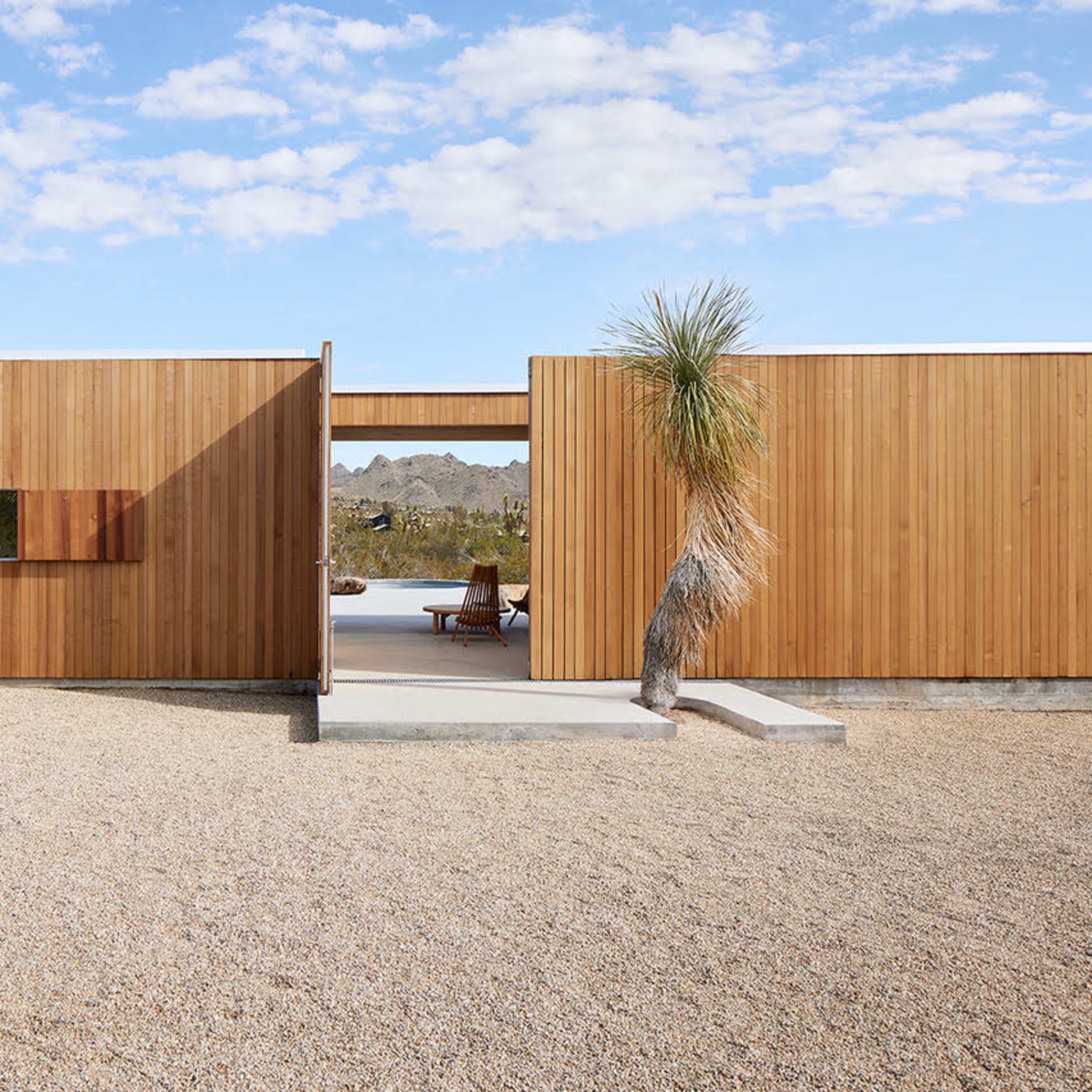 A modern wooden house surrounded by a gravel landscape and desert vegetation.