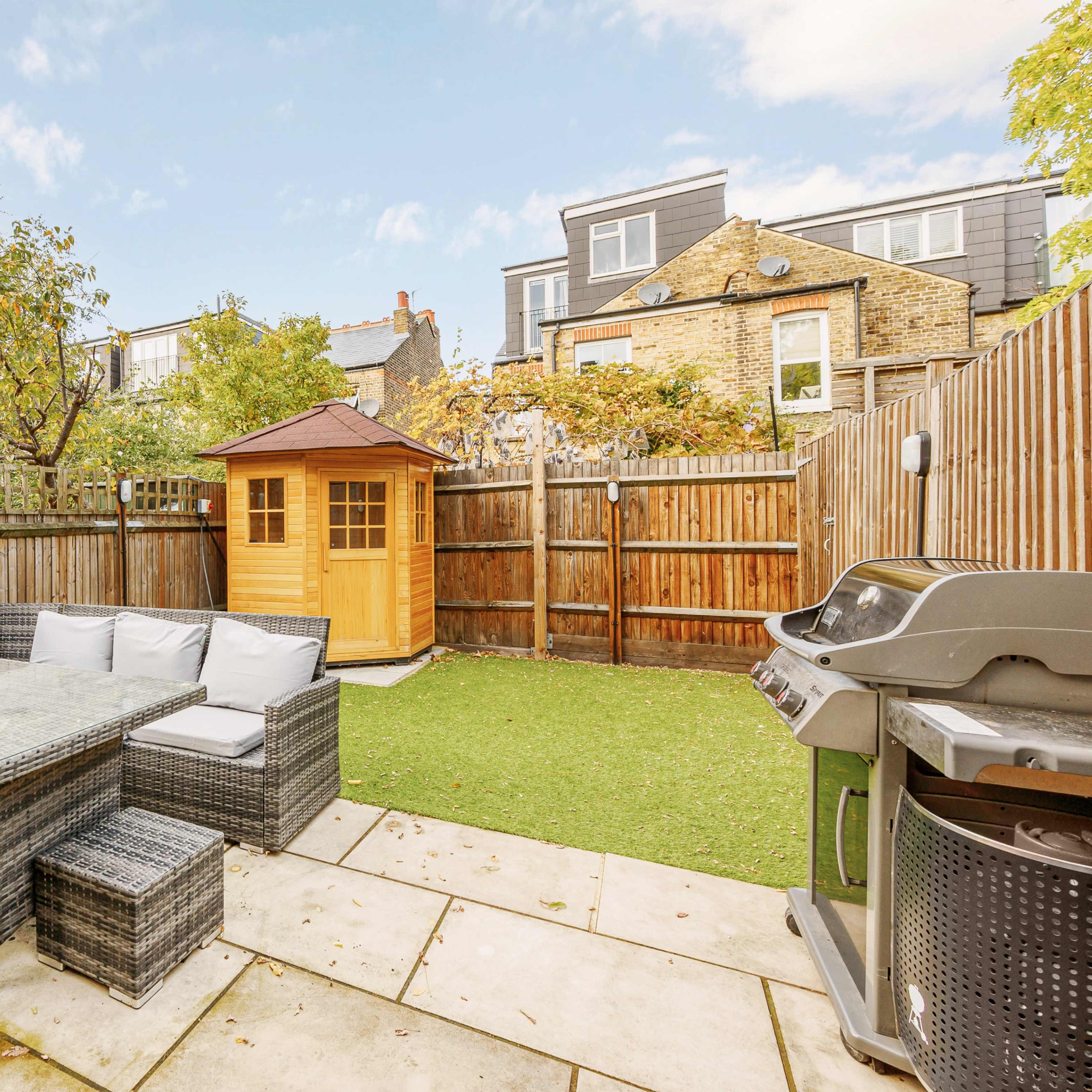 A backyard features a seating area with a glass-top table, a wooden shed, a barbecue grill, and a well-maintained lawn surrounded by wooden fencing.