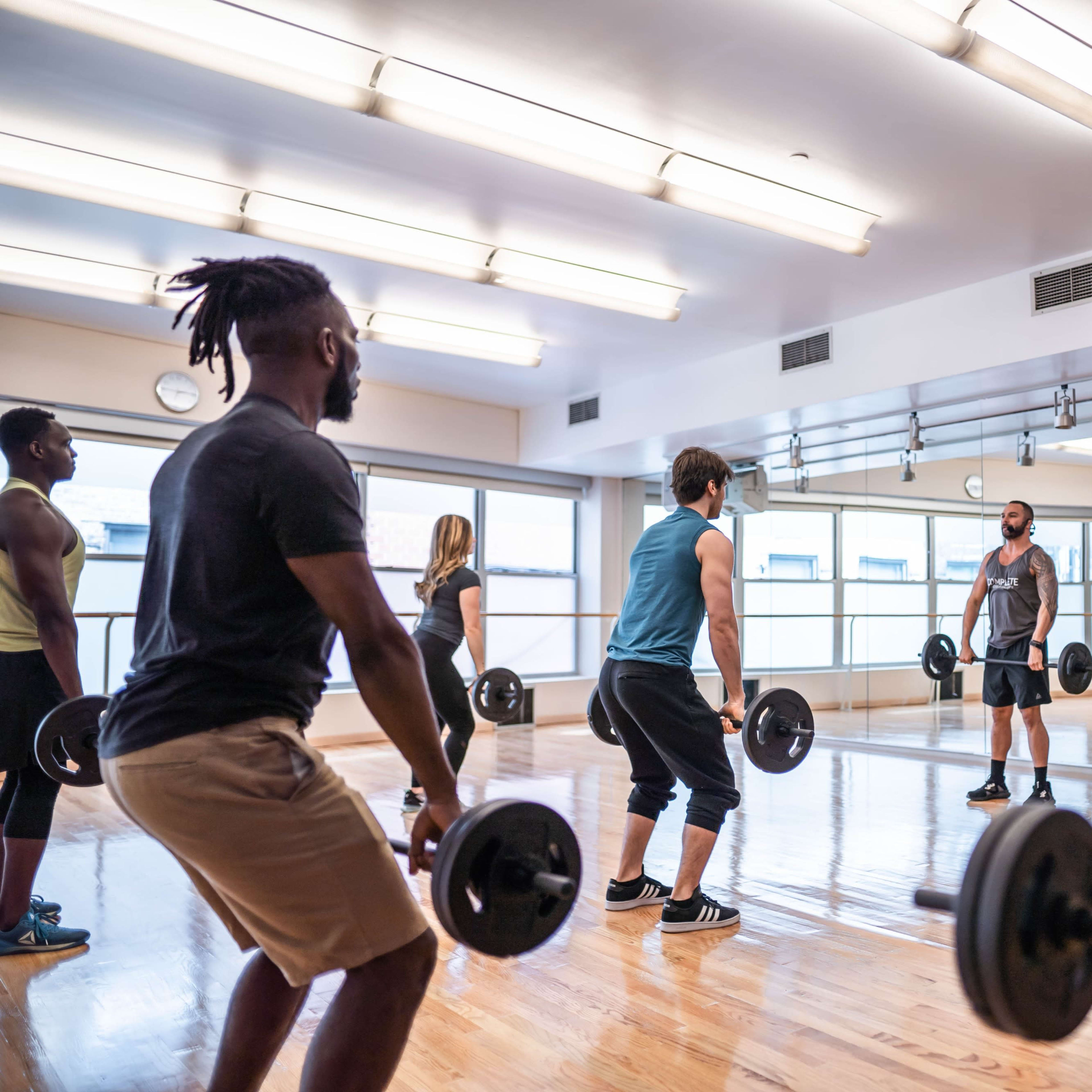 A group of people is lifting weights in a bright gym studio with mirrors and wooden floors.