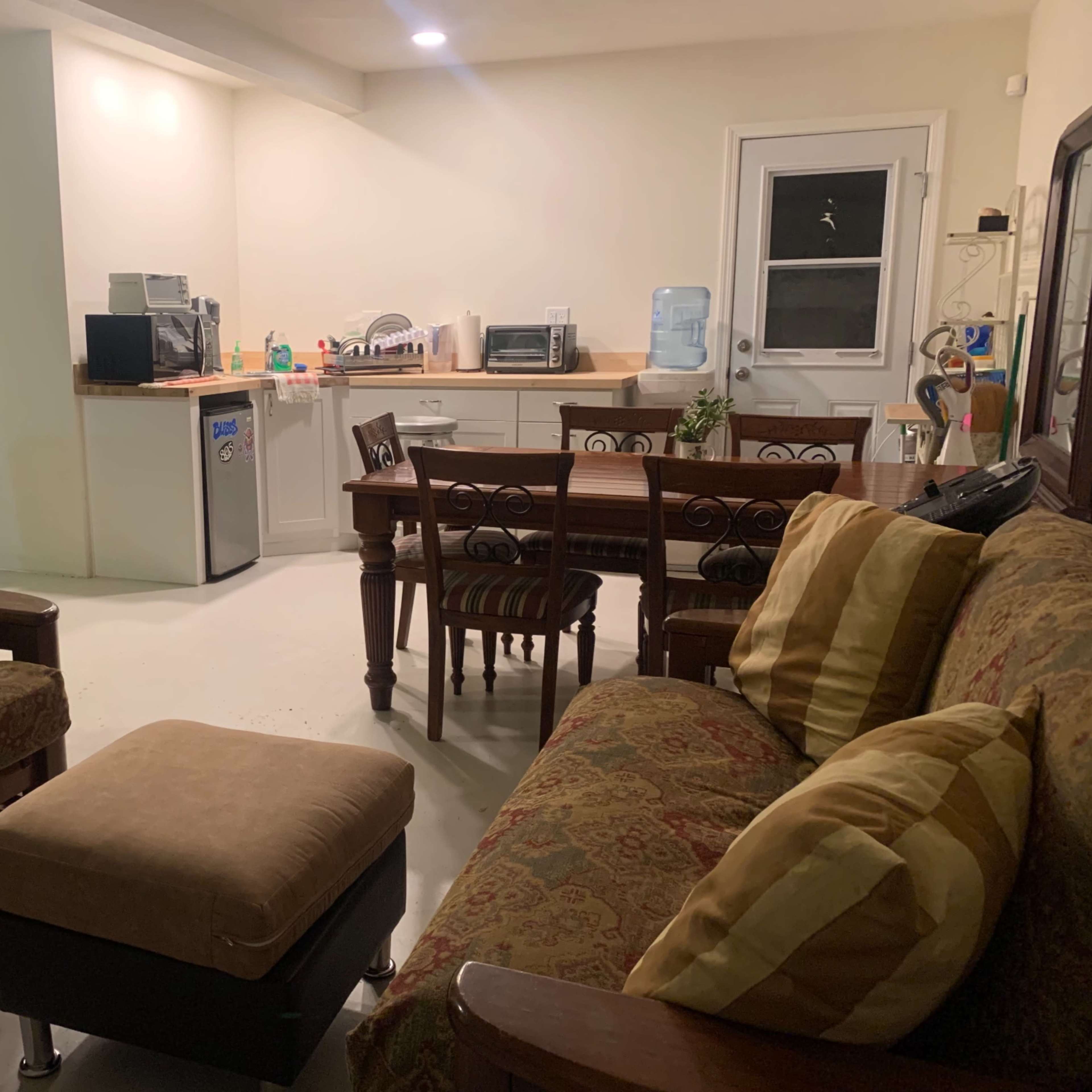 A living area with two couches, an ottoman, and a dining table near a kitchenette with a refrigerator and various appliances.