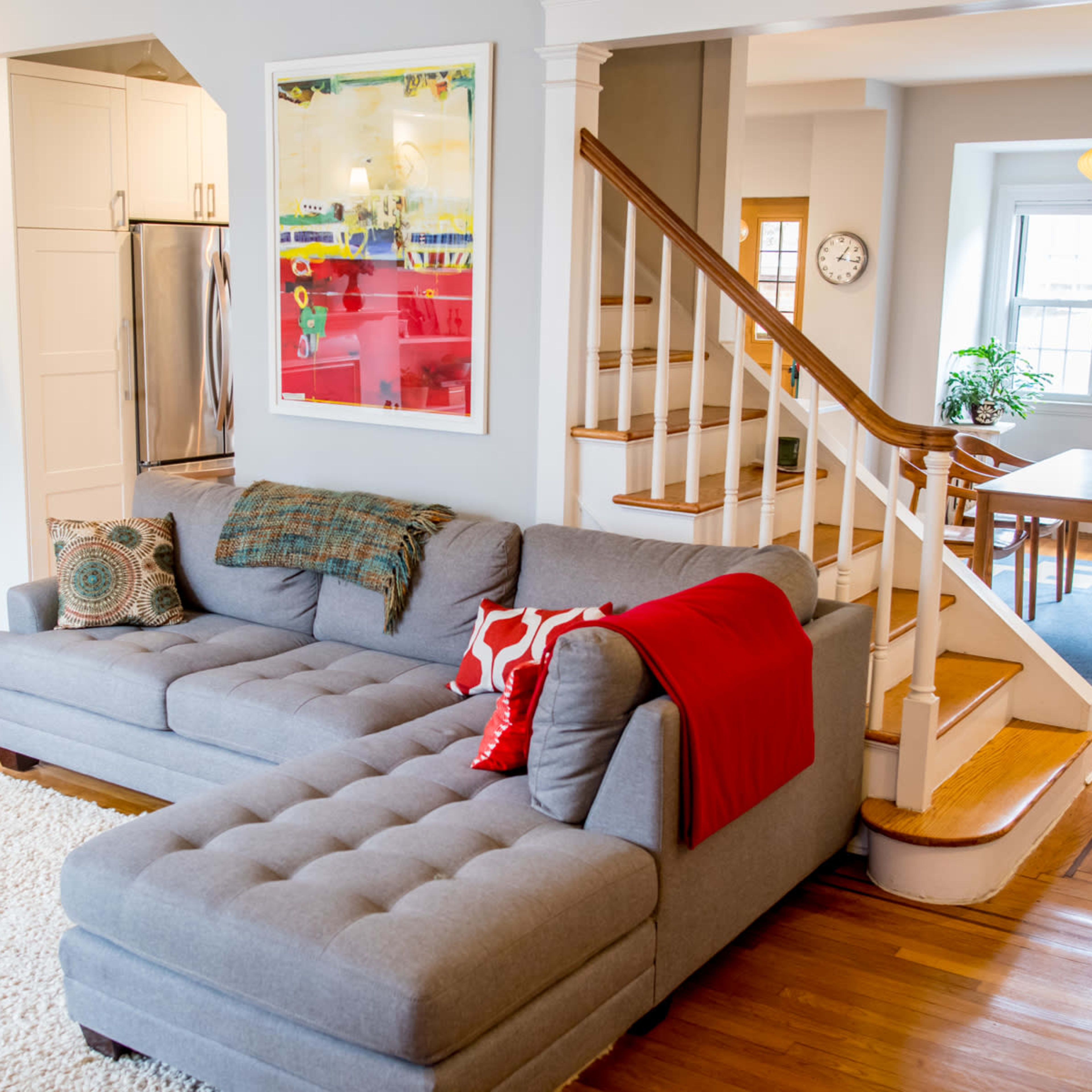 A living room with a gray sectional sofa, a red throw blanket, wooden stairs leading to an upper level, and a dining area in the background.