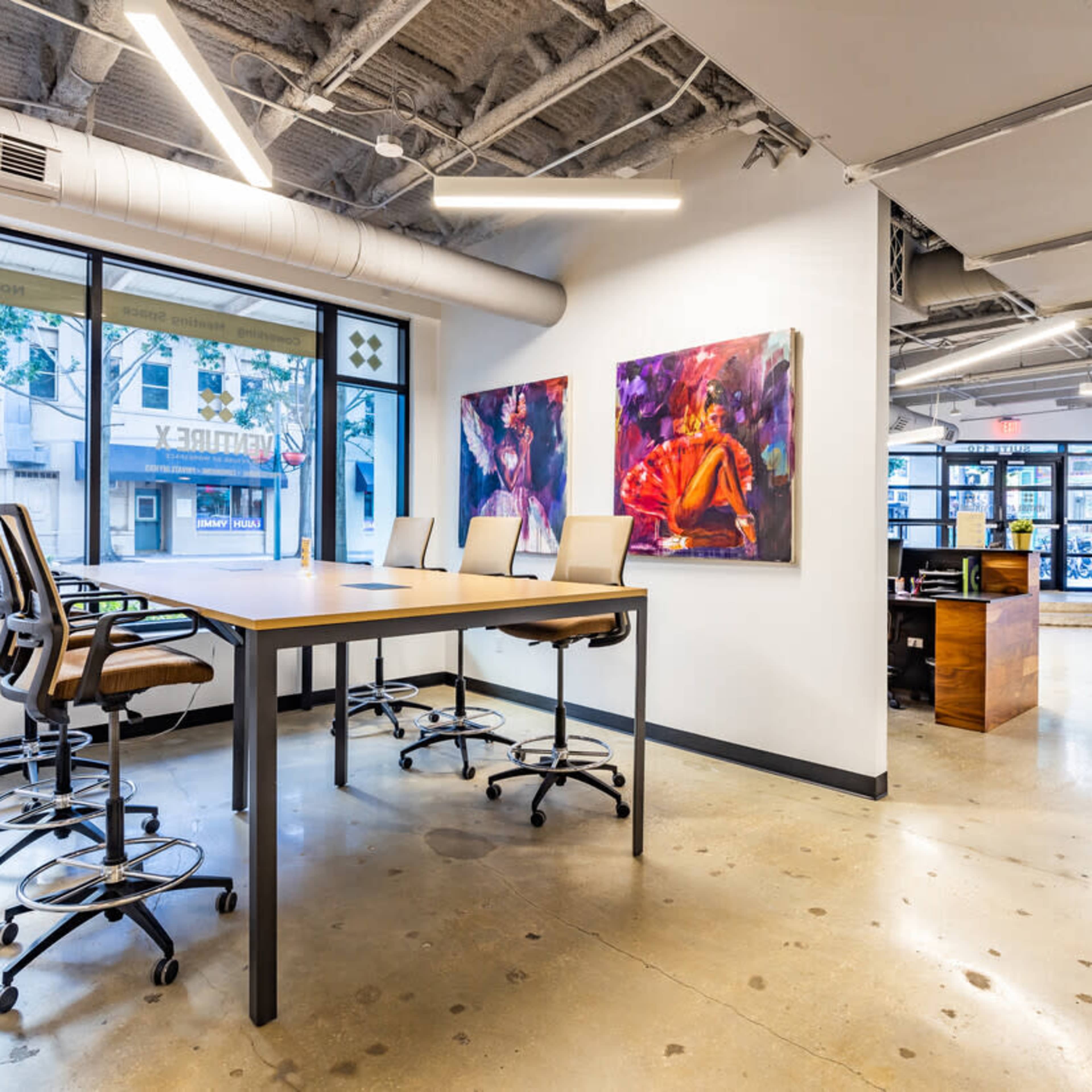A modern office space with a large, rectangular table surrounded by high-backed chairs, two colorful artworks on the wall, and large windows facing the street.